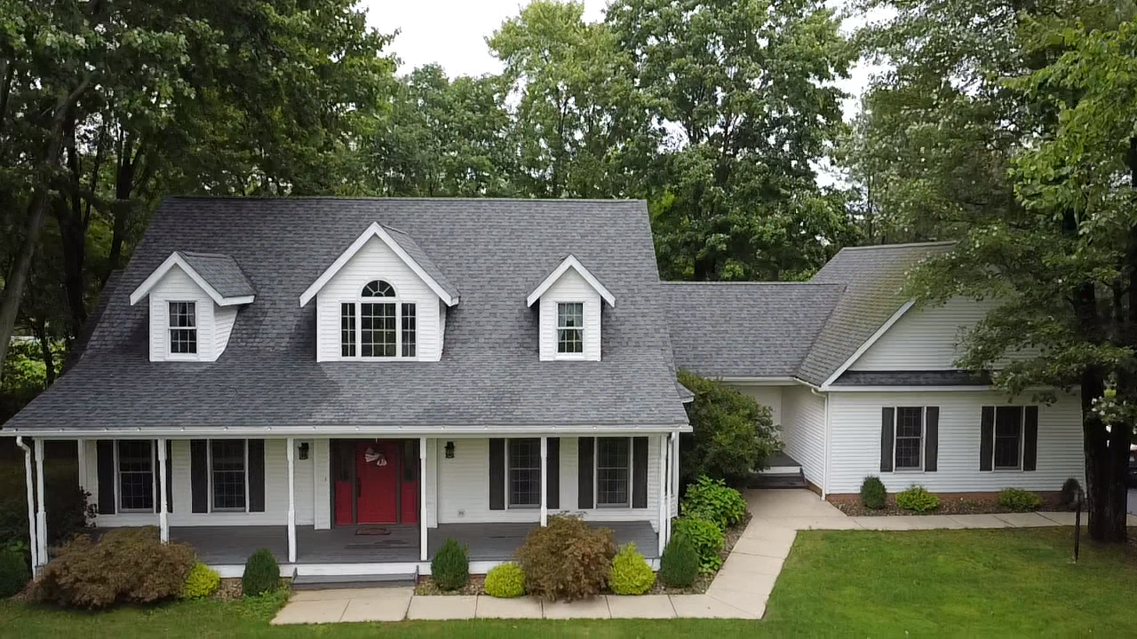 White Cape Cod home with red front door, gray roof, dormers, covered porch, and lush green trees in background.