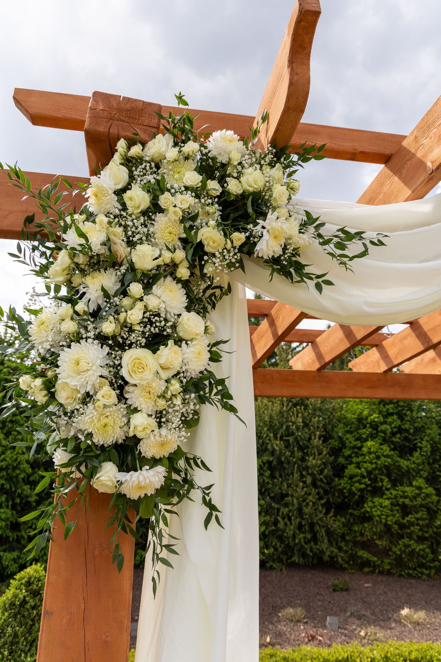 White floral arrangement with roses and carnations on a wooden wedding arch draped with white fabric outdoors.