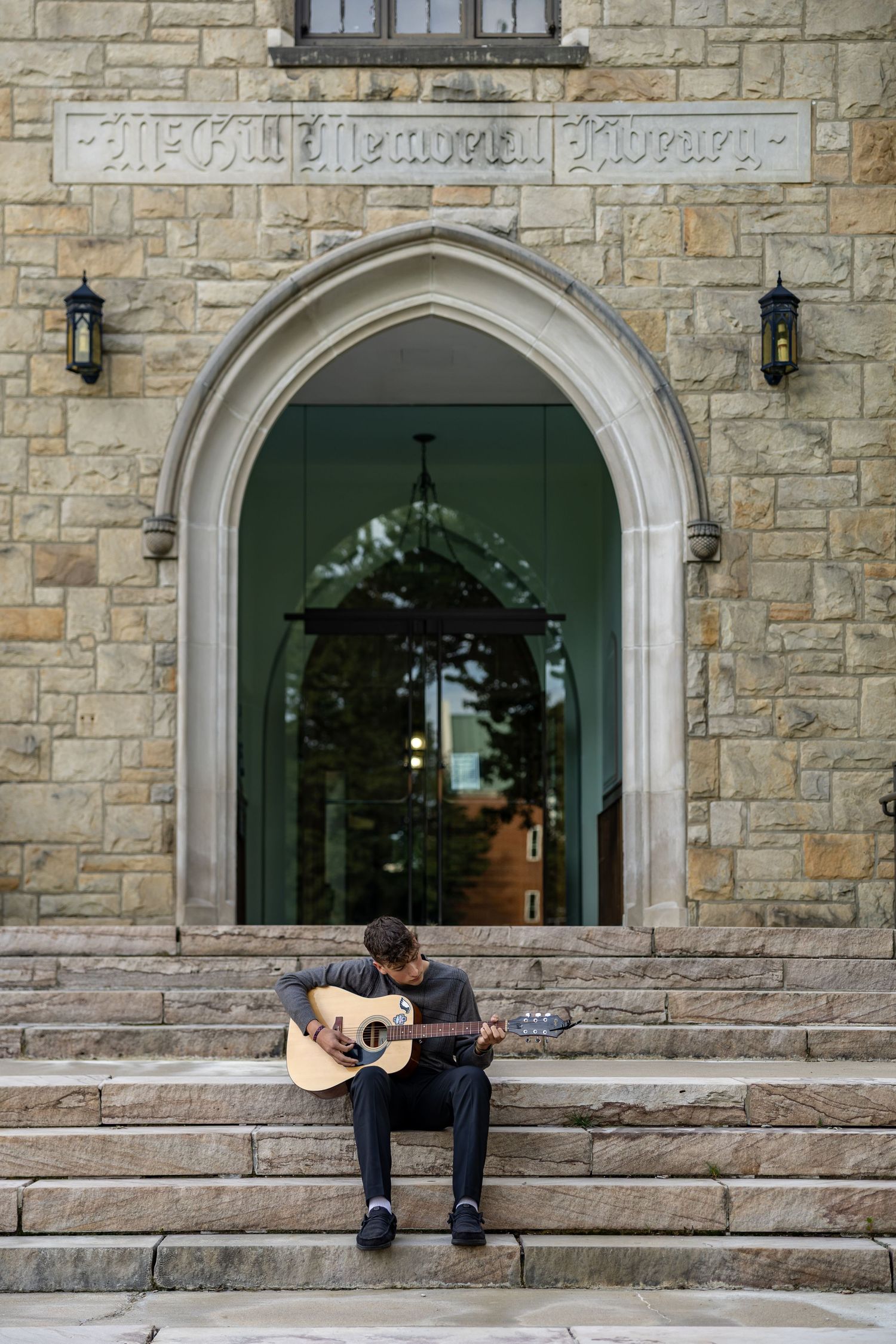 A musician plays acoustic guitar on stone steps outside McGill Memorial Library's Gothic arch entrance.