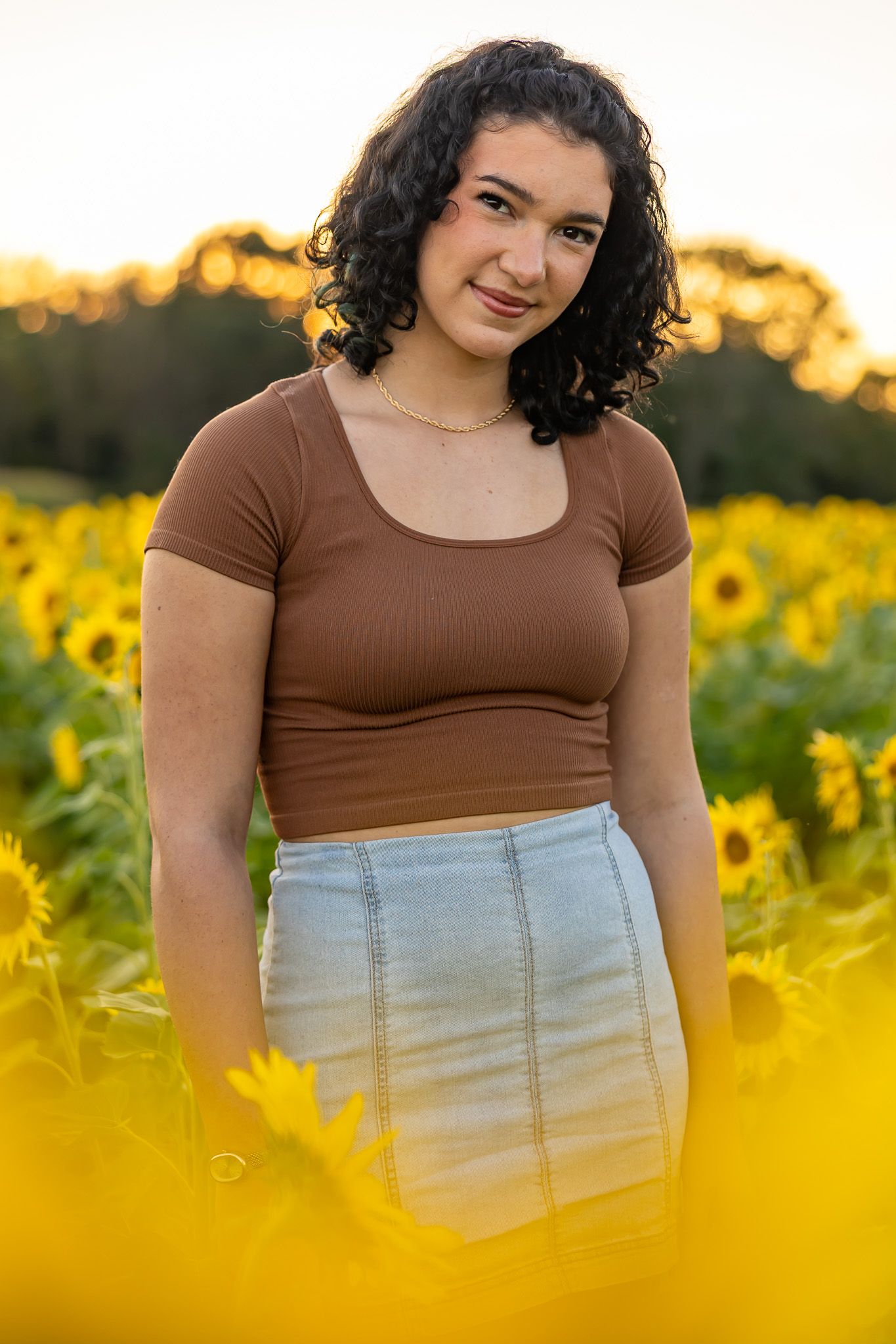 Young woman with curly hair in brown crop top and denim skirt posing in a vibrant sunflower field at golden hour.