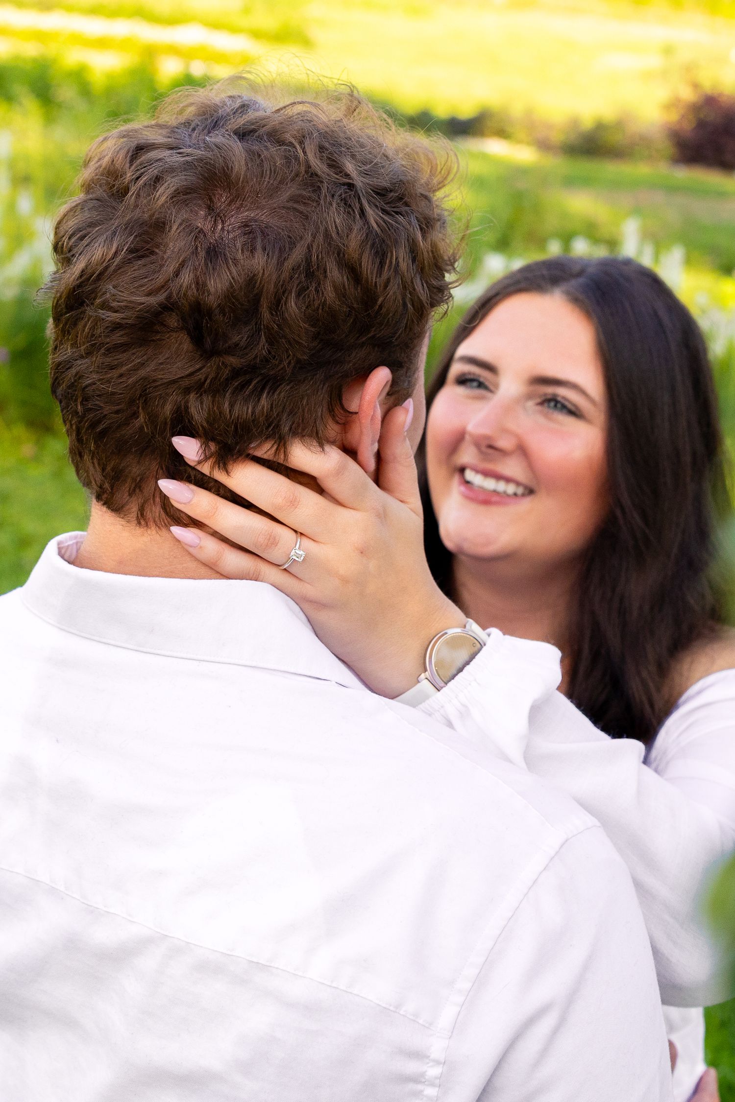 Smiling bride-to-be embraces her fiance outdoors, showing off her engagement ring in a sunny green field.