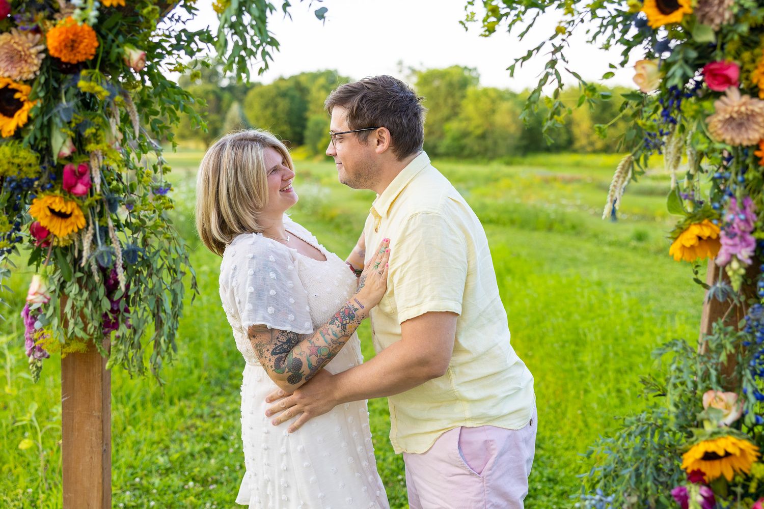 Couple embracing under a colorful floral arch in a green meadow during an outdoor ceremony.