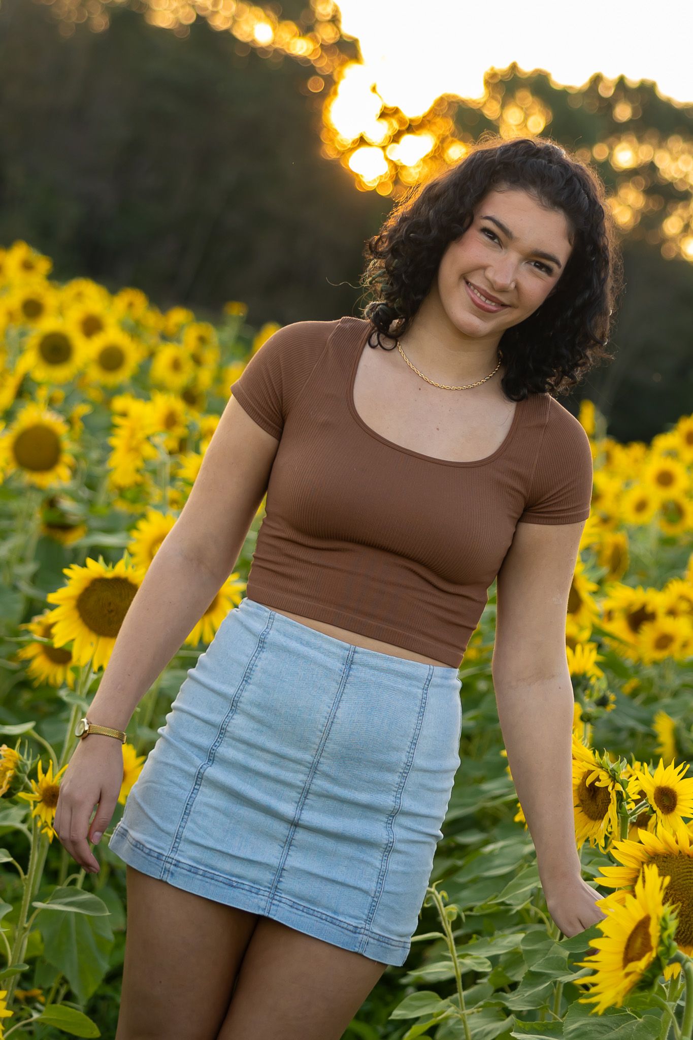 Young woman in brown crop top and denim skirt smiling in a sunflower field at golden hour sunset.