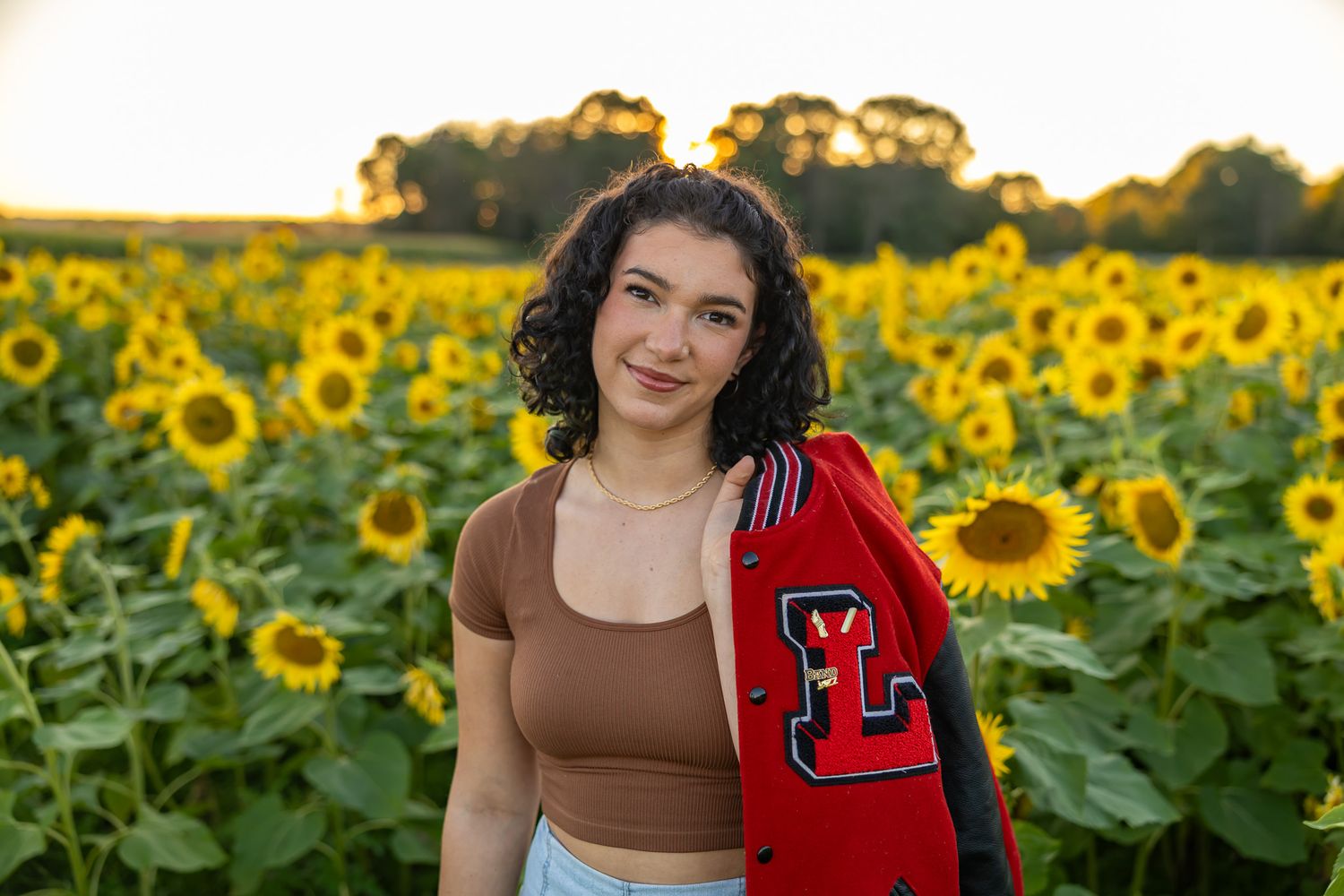 Teen girl in brown crop top holds red varsity jacket in a sunny sunflower field at golden hour.