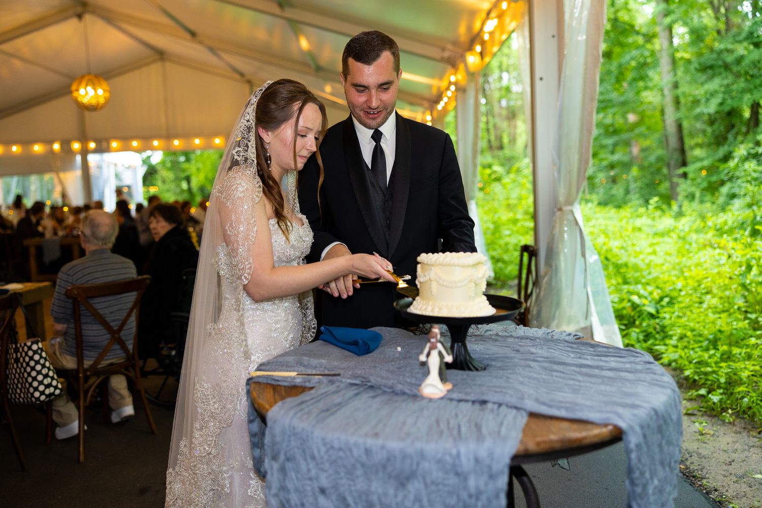 Bride and groom cutting white wedding cake under elegant tent with string lights and lush green trees in background.