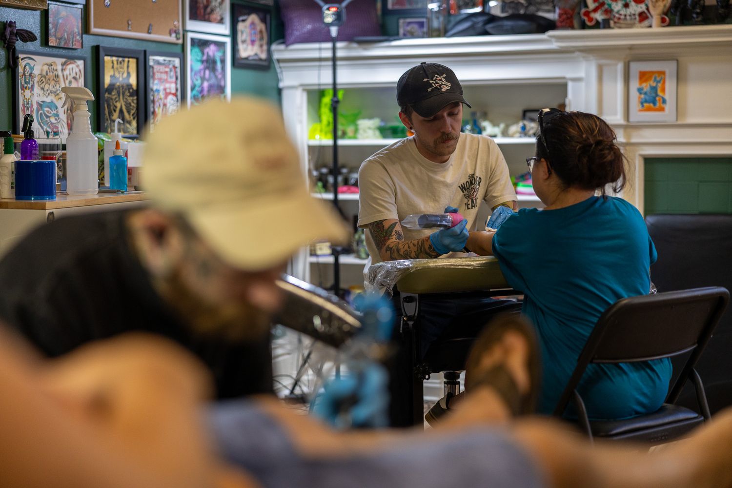 Tattoo artist in white shirt working on a client's arm inside a busy tattoo parlor.
