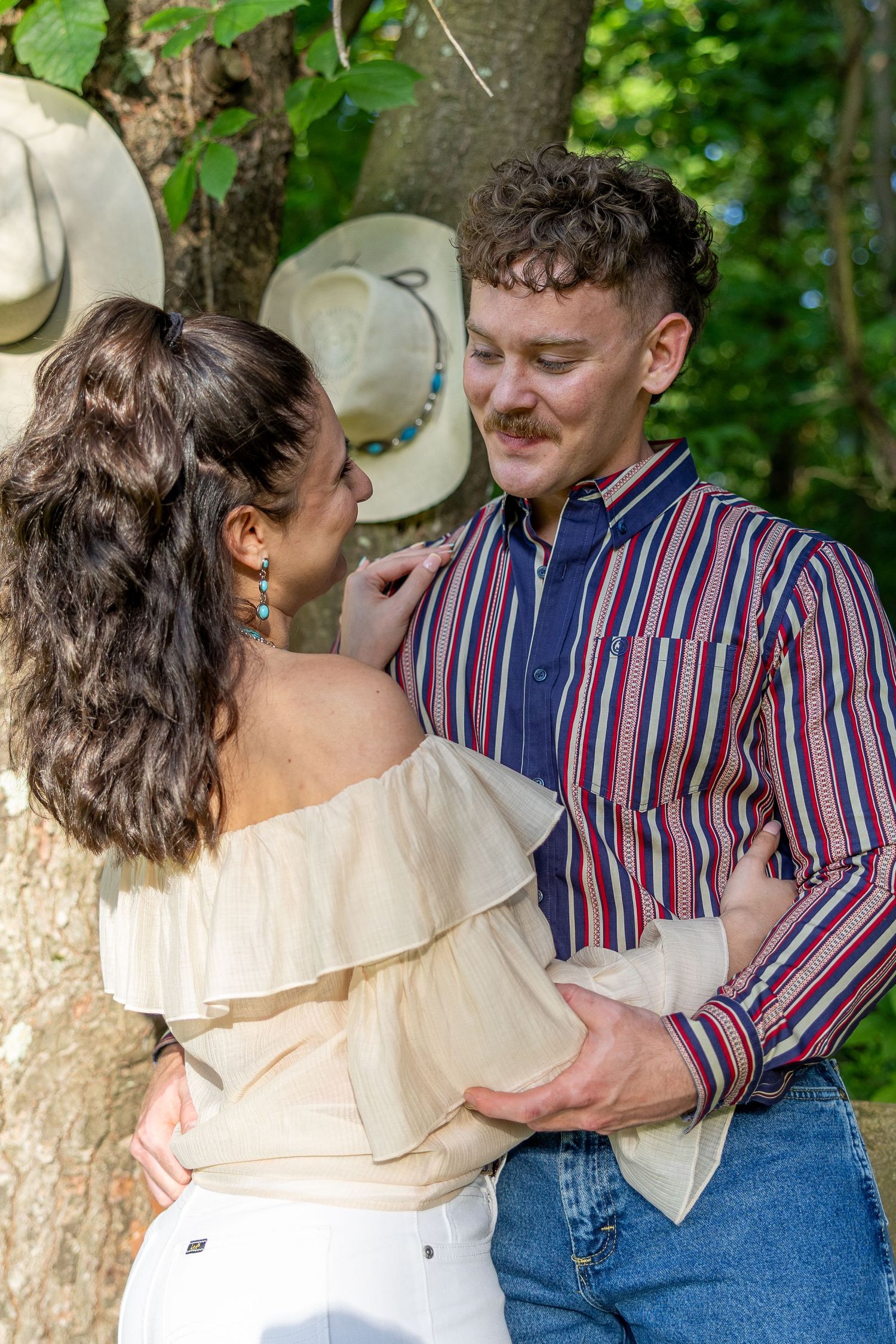 Couple embracing near tree with straw hats hanging, man in striped shirt and jeans.