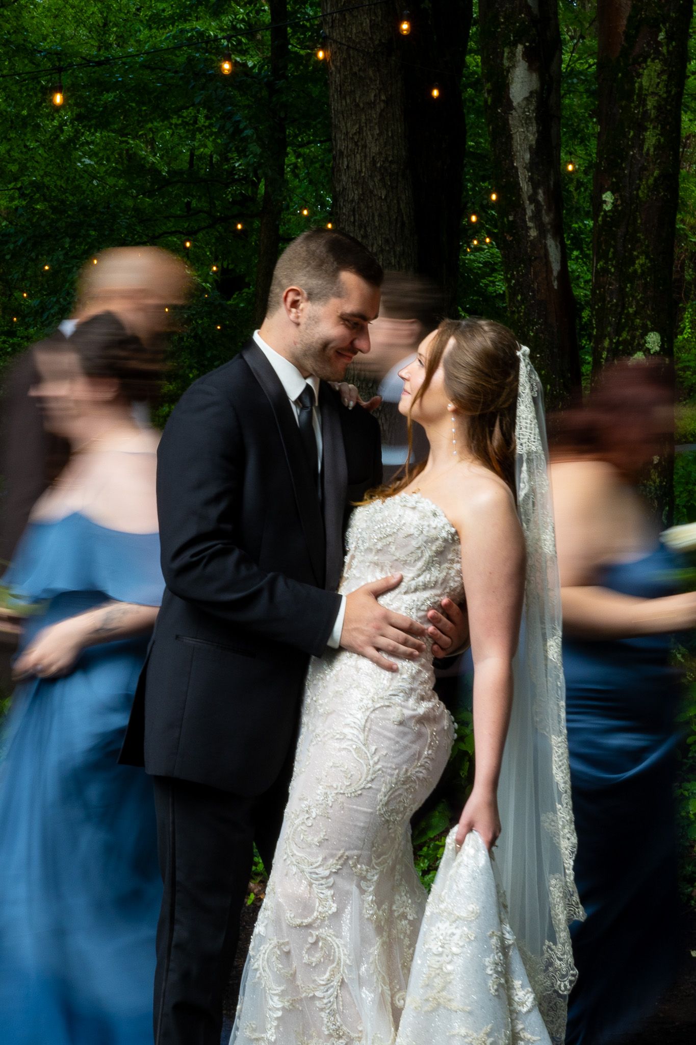 Bride and groom share romantic moment surrounded by blurred bridesmaids in blue in forest setting.