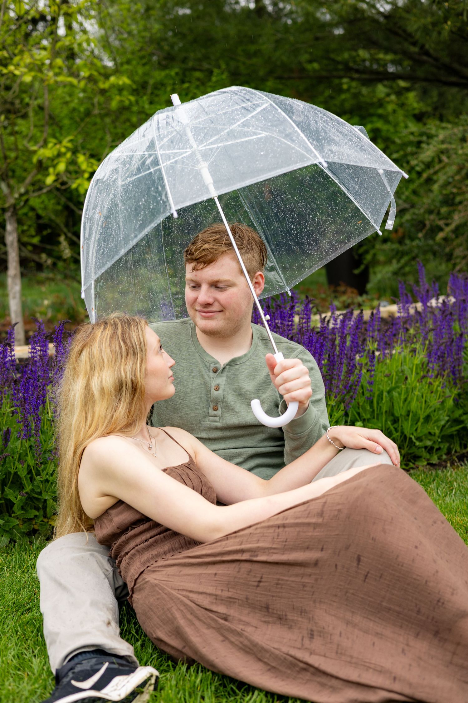 Couple relaxing on grass under a clear umbrella surrounded by purple flowers.
