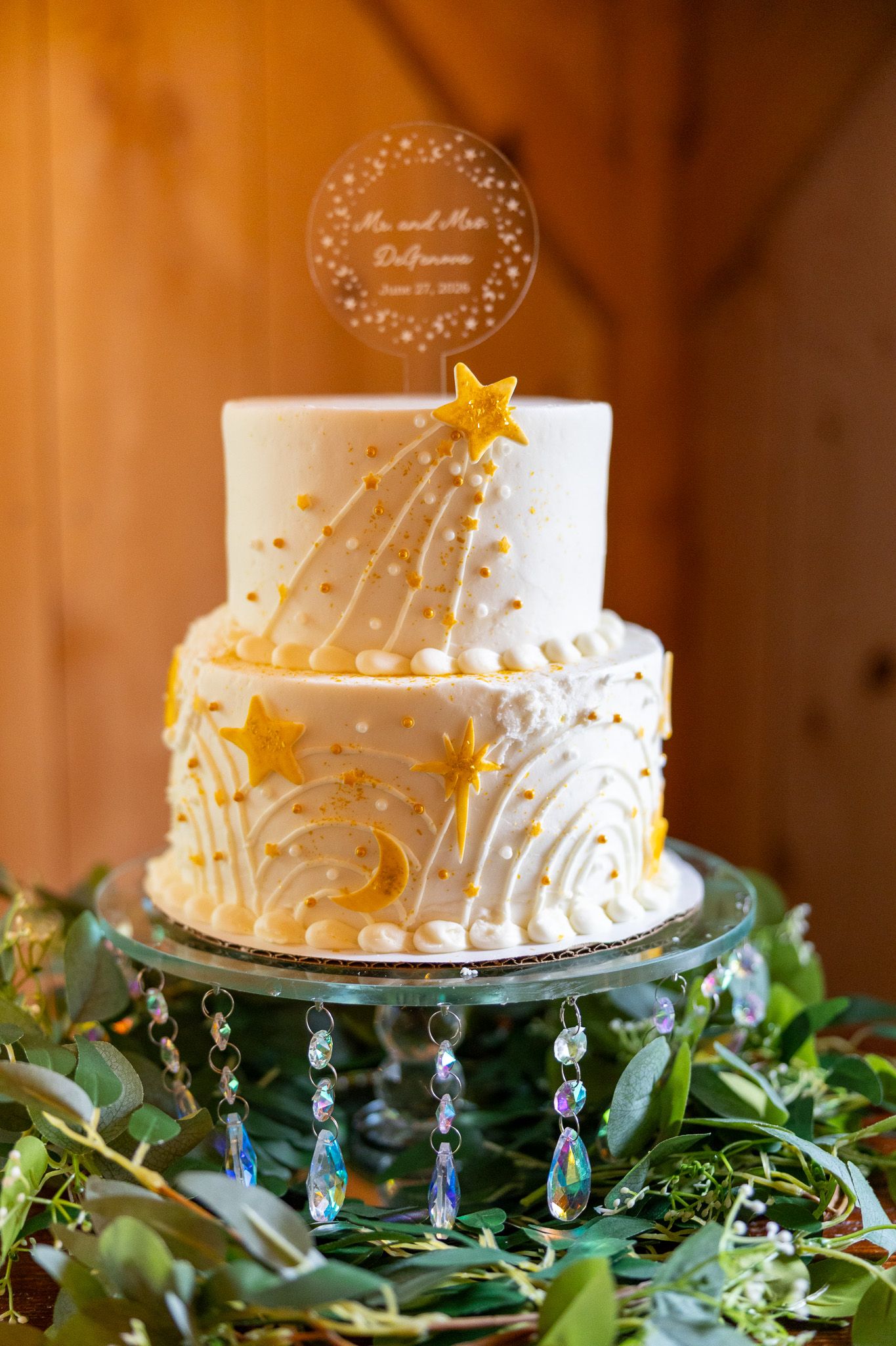 Two-tier white wedding cake with gold stars, moon, and swirl decorations on a crystal cake stand surrounded by greenery.