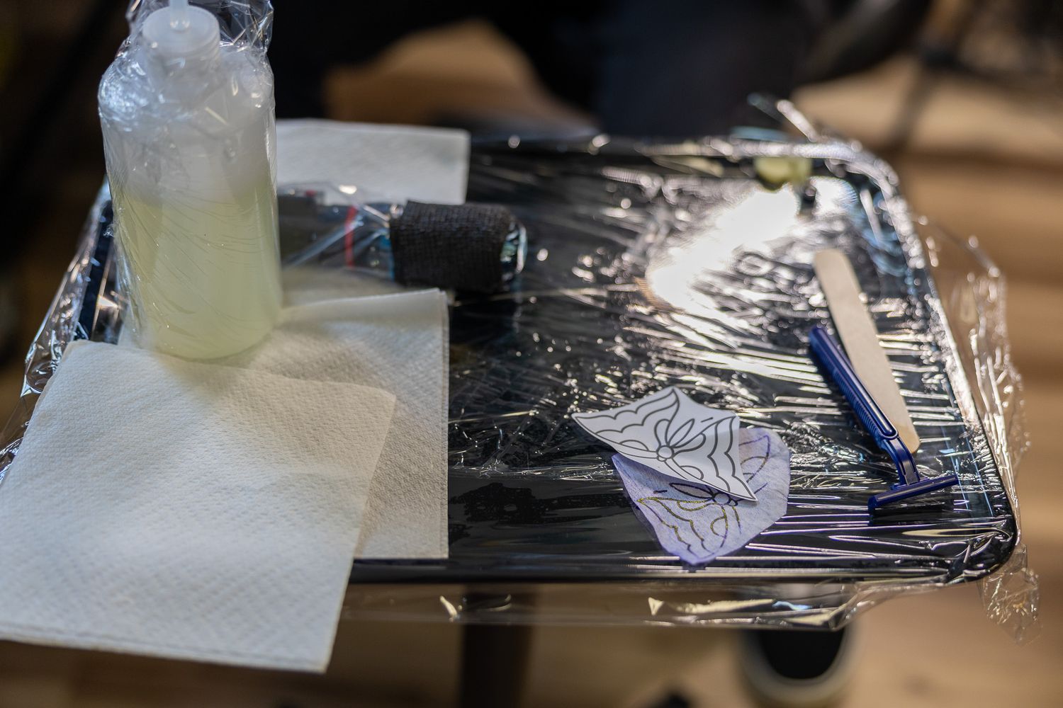 Tattoo artist preparation tray wrapped in plastic with supplies including a razor, stencil, and spray bottle.