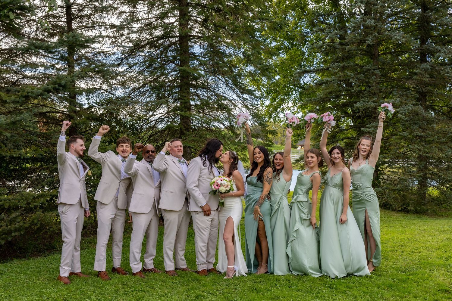 Wedding party celebrating outdoors in green bridesmaid dresses and gray suits, raising arms joyfully among tall trees.