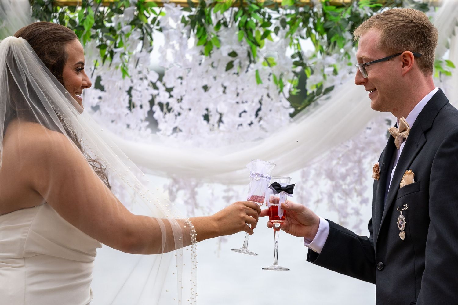 Bride and groom toasting with champagne glasses at wedding ceremony under floral arch.