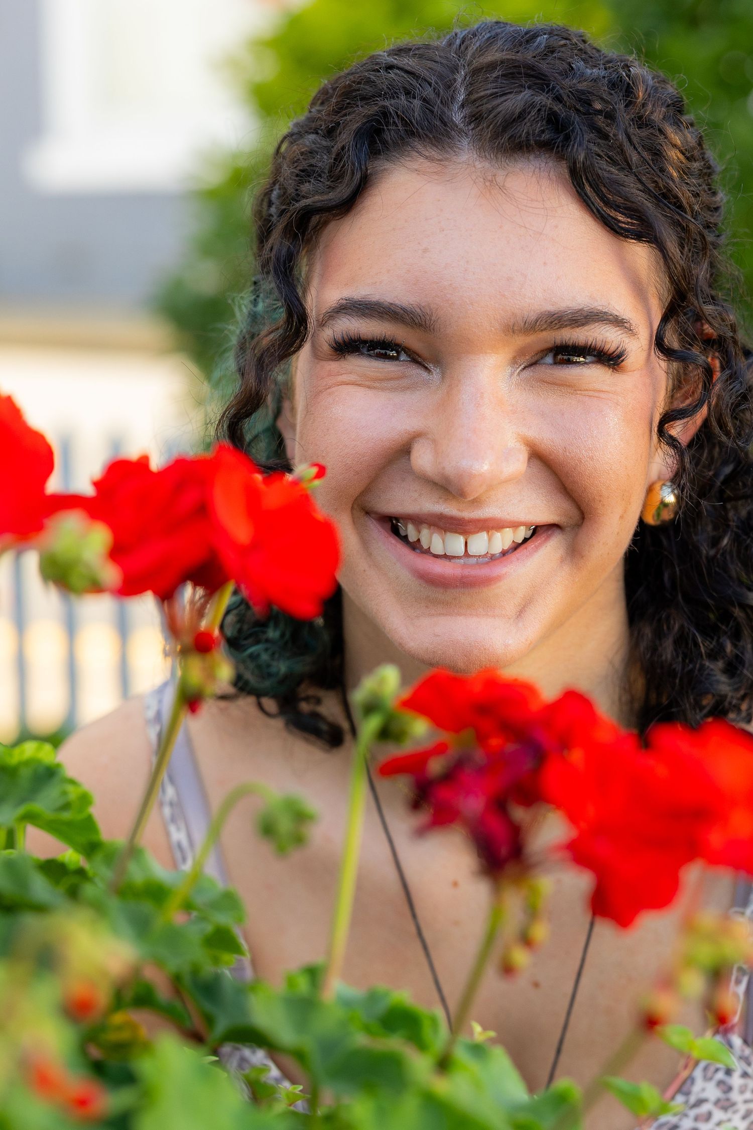 Smiling young woman with curly hair surrounded by vibrant red geranium flowers outdoors.