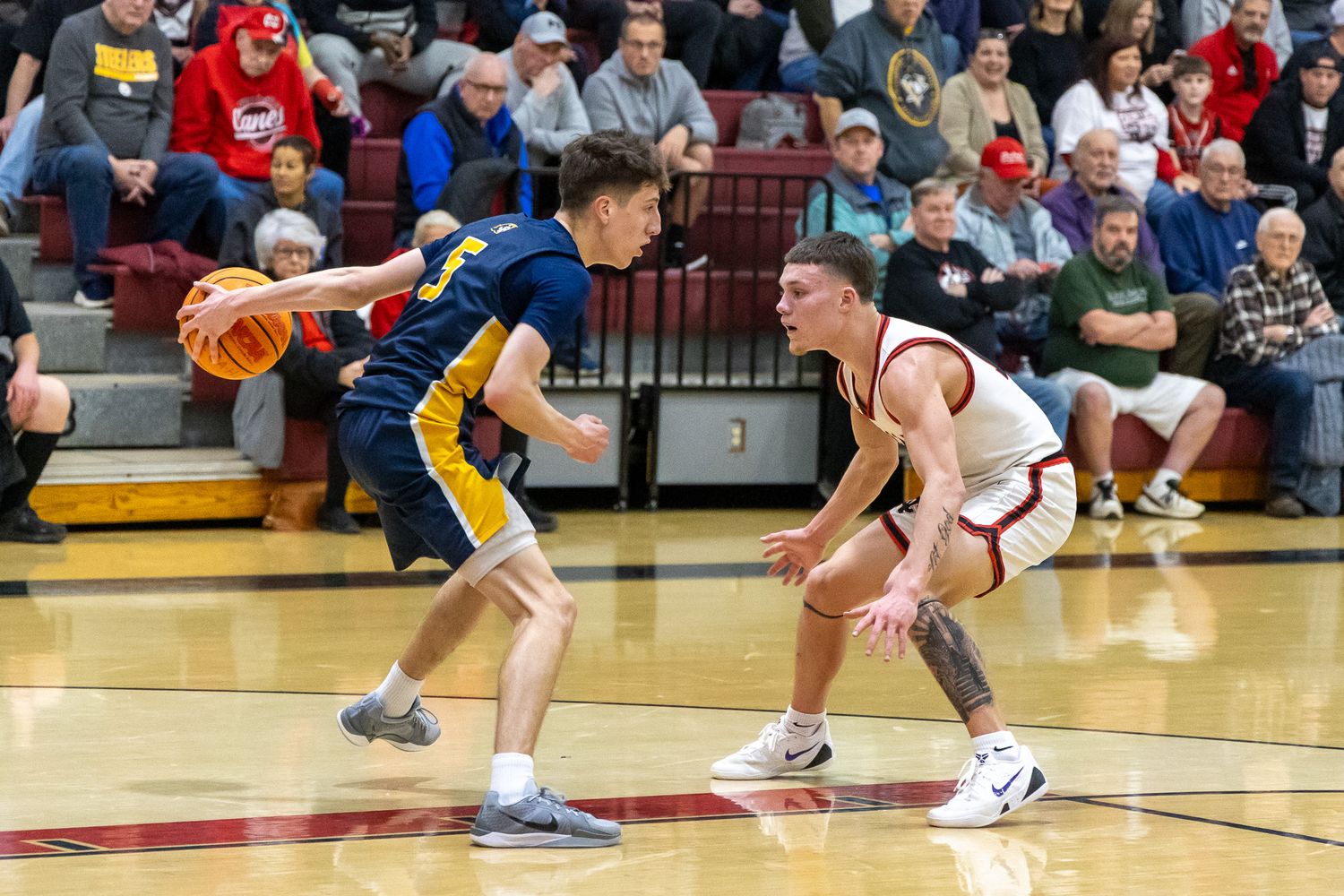 Male basketball player dribbles ball while defender guards closely in crowded gymnasium.