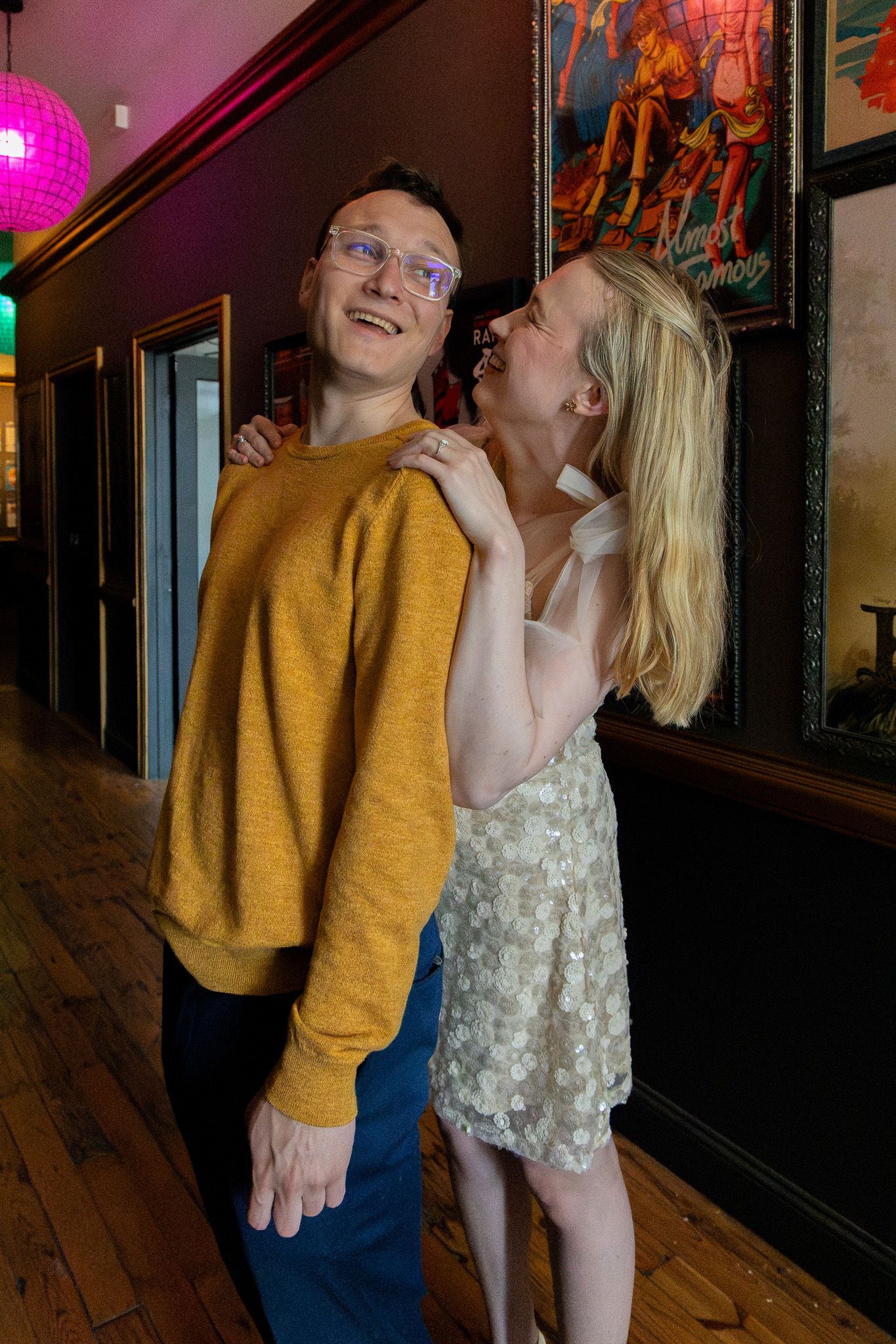 Playful couple posing in decorated hallway, woman in floral dress embracing man in mustard sweater.