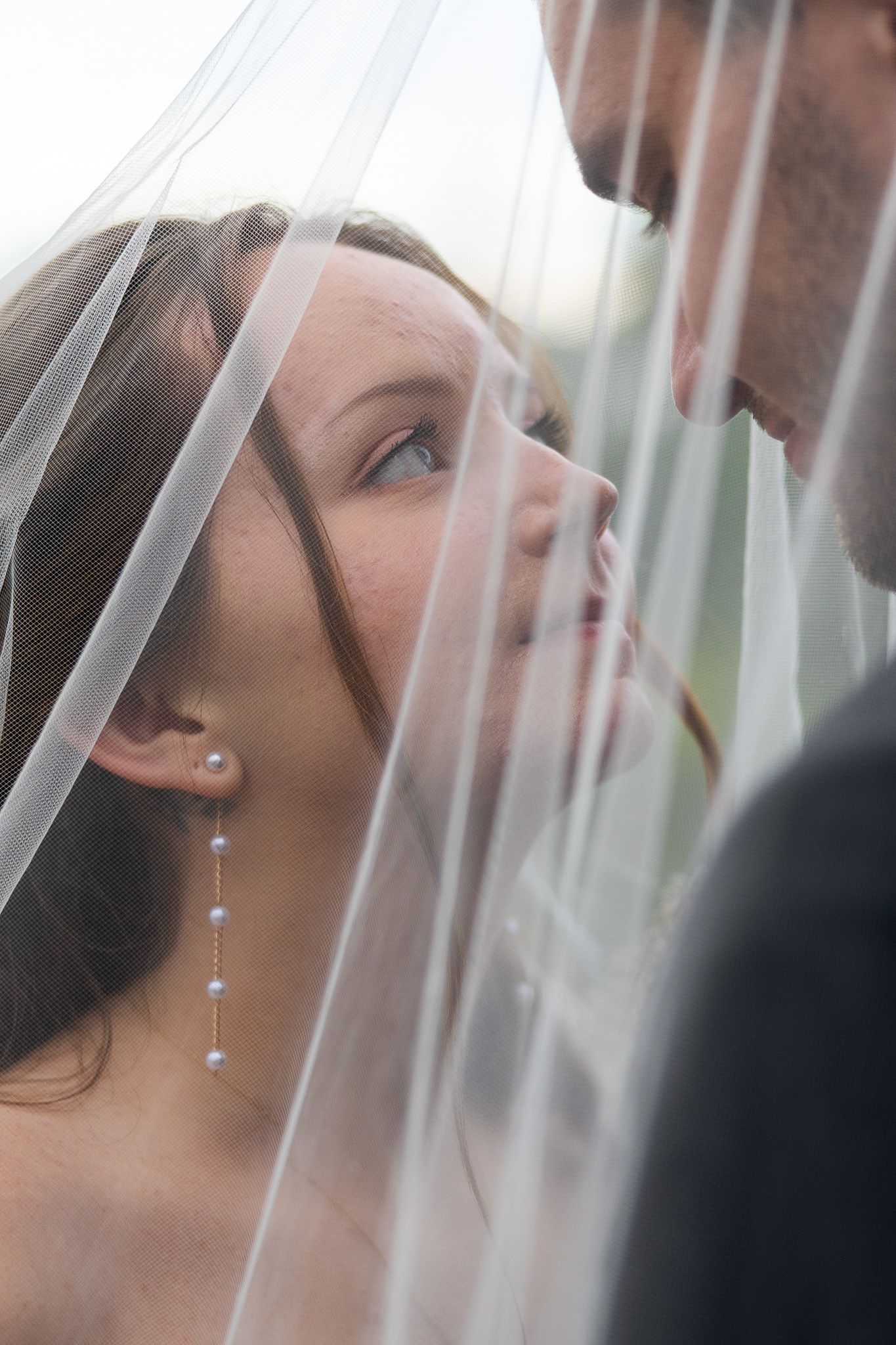 Bride and groom share an intimate moment under a wedding veil, bride gazing up with pearl drop earrings.