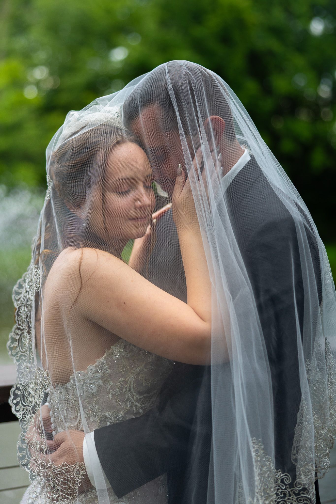 Bride and groom share an intimate moment under a lace wedding veil outdoors with green trees in the background.