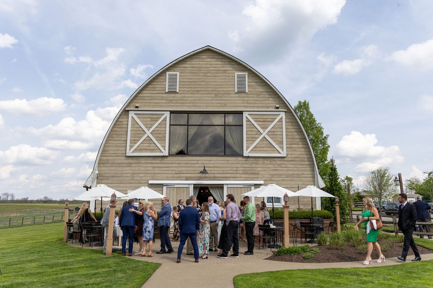 Guests mingle outside a rustic barn wedding venue with white umbrellas on a sunny day.