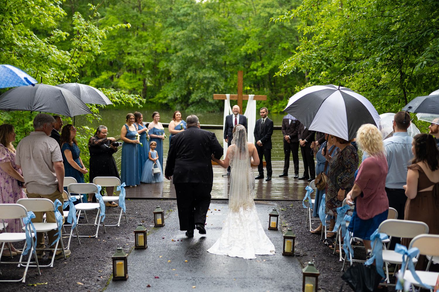 Bride walks down rainy outdoor aisle lined with lanterns and umbrella-holding guests toward wooden cross altar.
