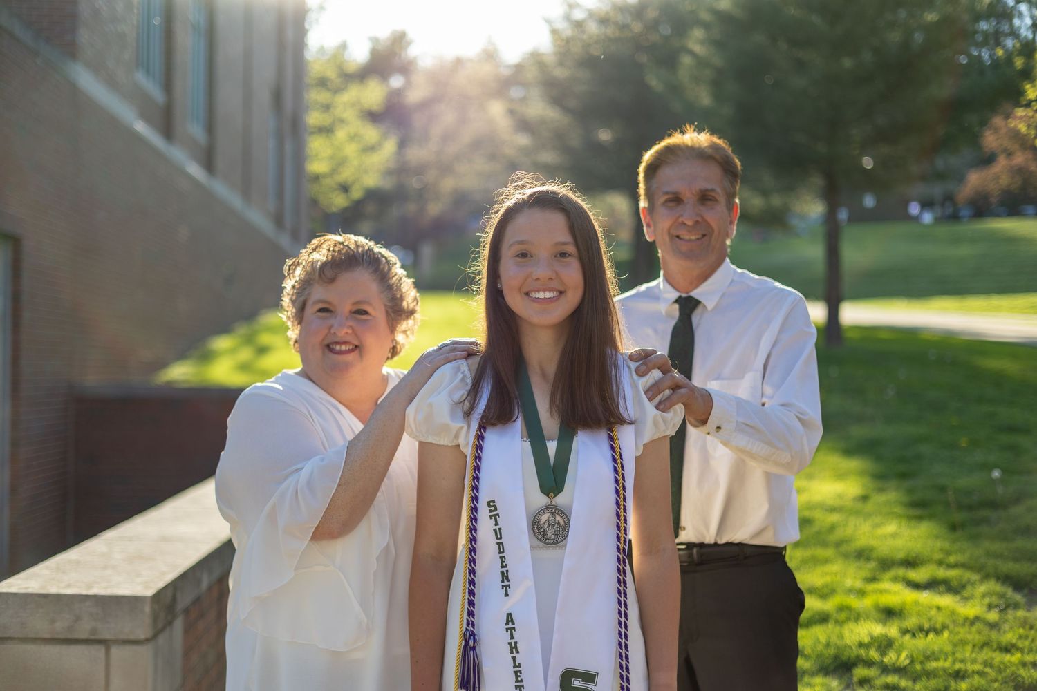 Female graduate with medal and cords poses smiling with two family members outside university building.
