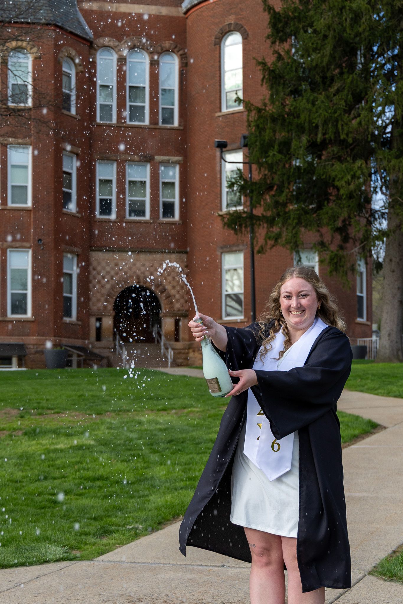 Female graduate in cap and gown pops champagne bottle outside Slippery Rock University building.