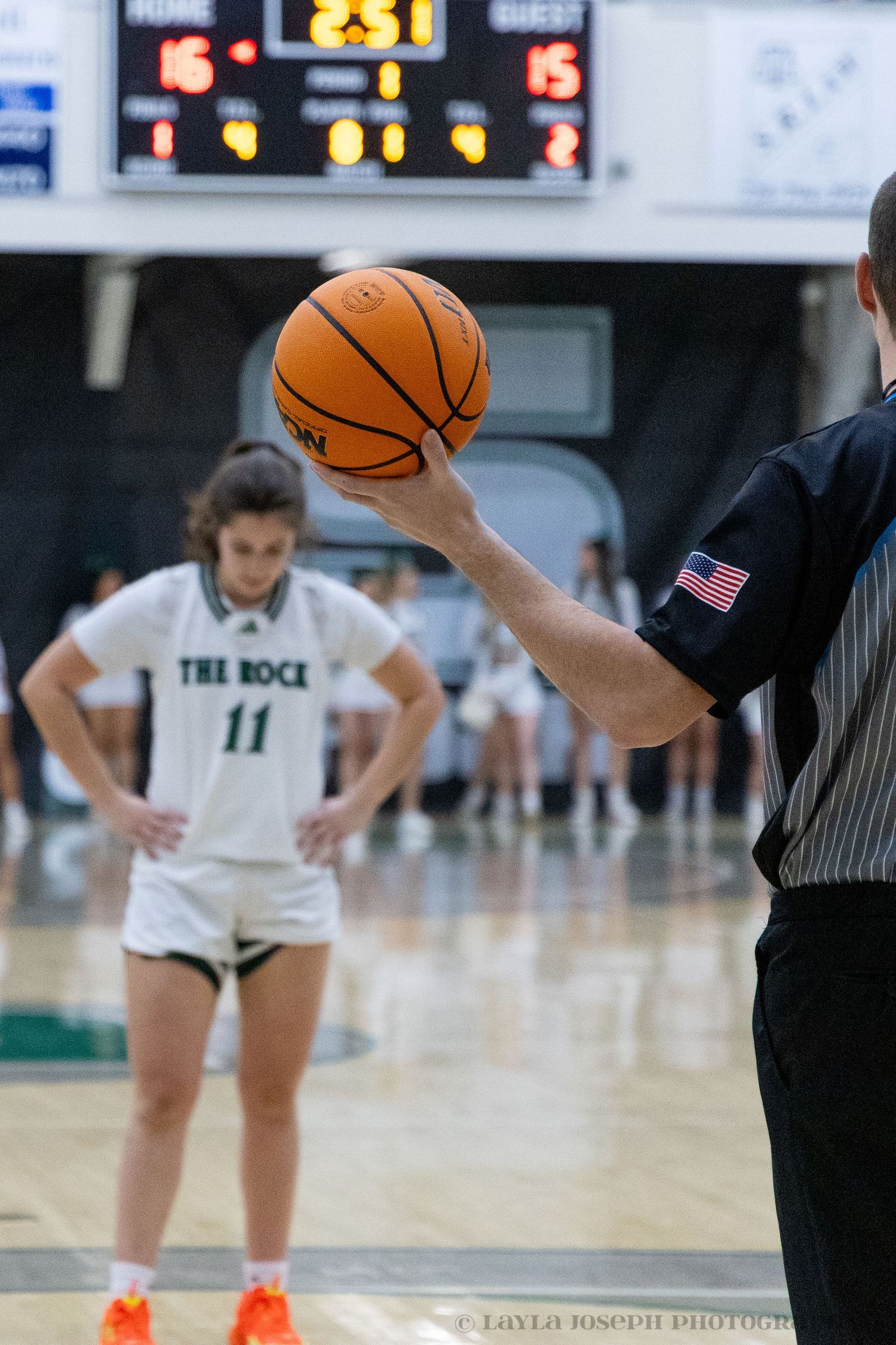 Basketball referee holds up ball near player number 11 in The Rock uniform during game.
