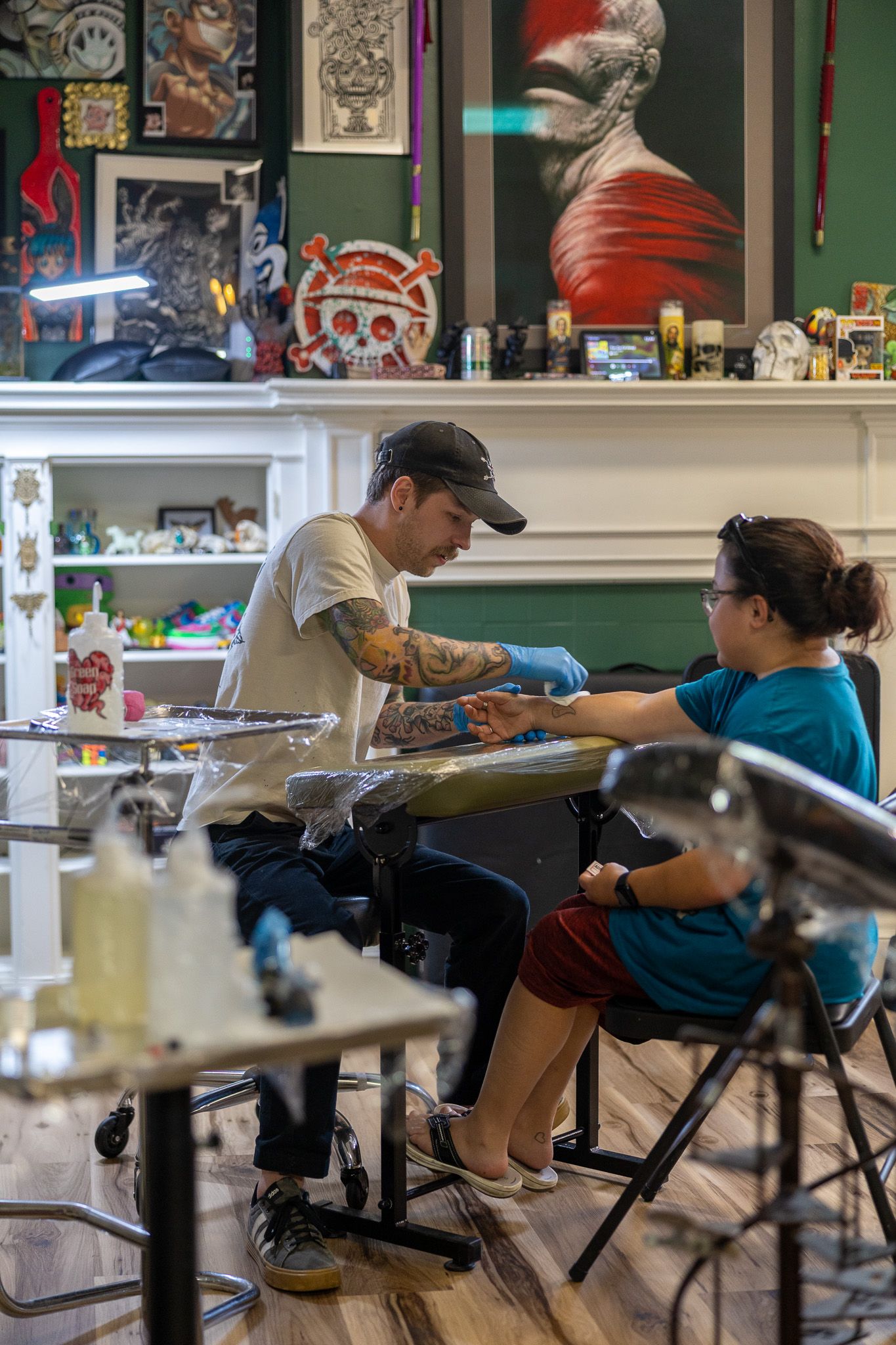 Tattoo artist in cap and gloves tattooing a client's hand in a decorated tattoo studio.