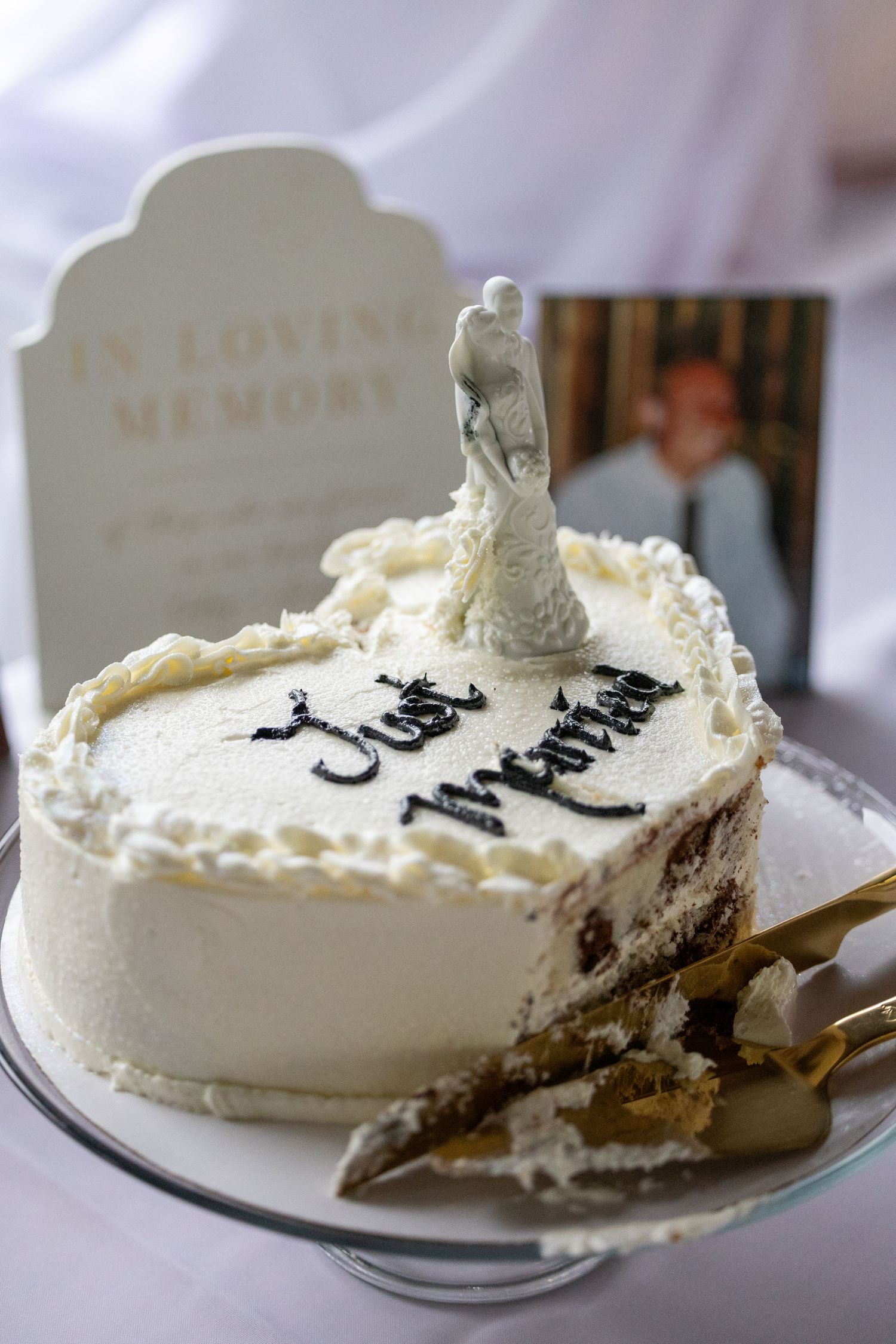 Wedding cake with 'Just Married' script, bride and groom topper, beside an 'In Loving Memory' tombstone decoration.