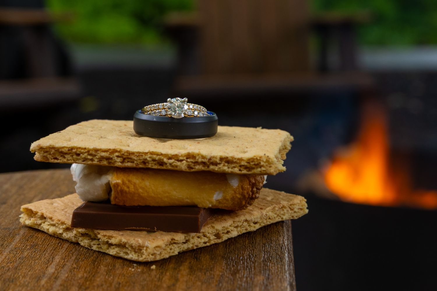 Two wedding rings rest on a s'more with graham crackers, chocolate, and marshmallow, with a campfire in the background.