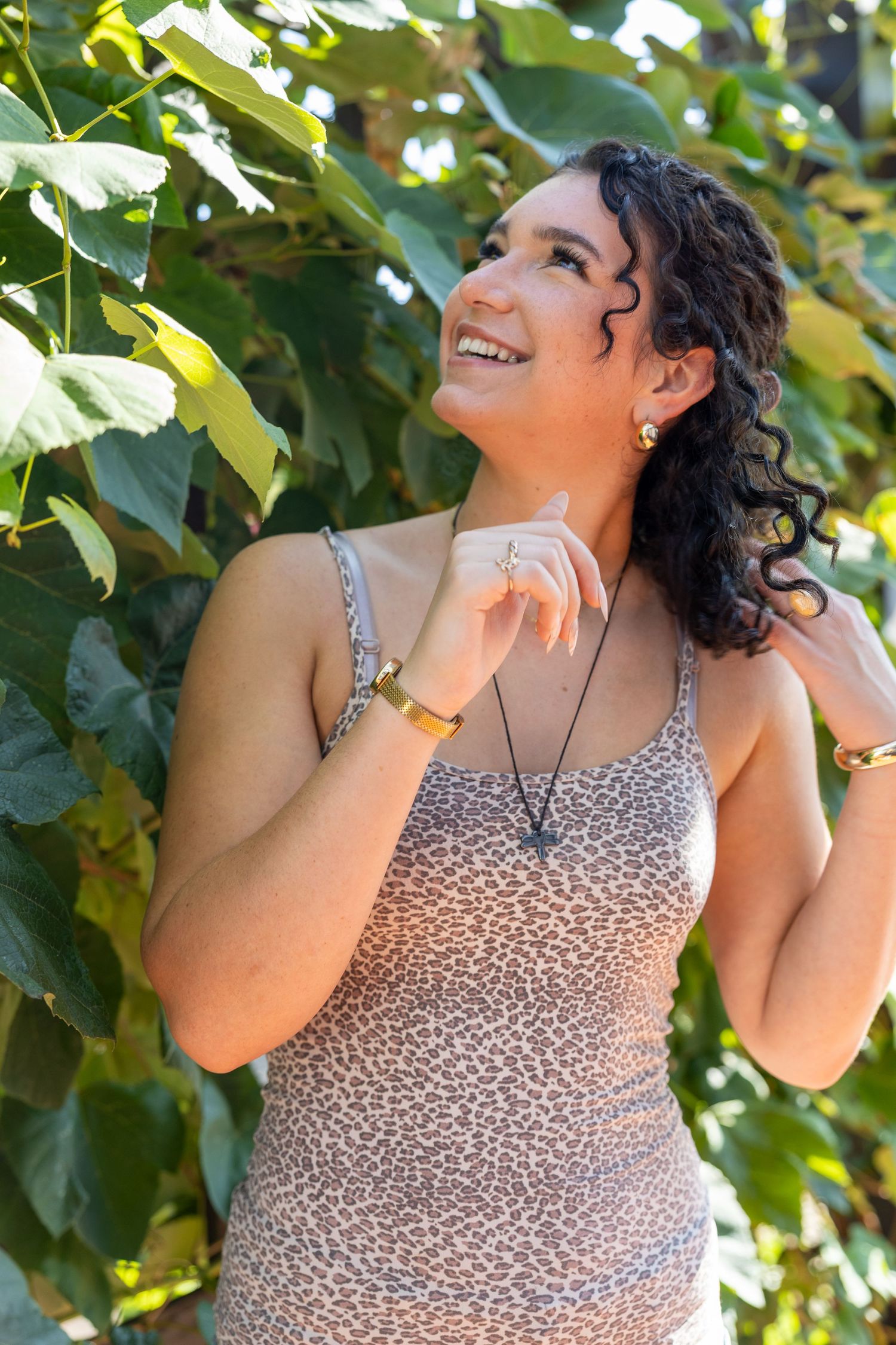 Smiling woman in leopard print dress with curly hair, gold jewelry, posing outdoors among lush green leaves.