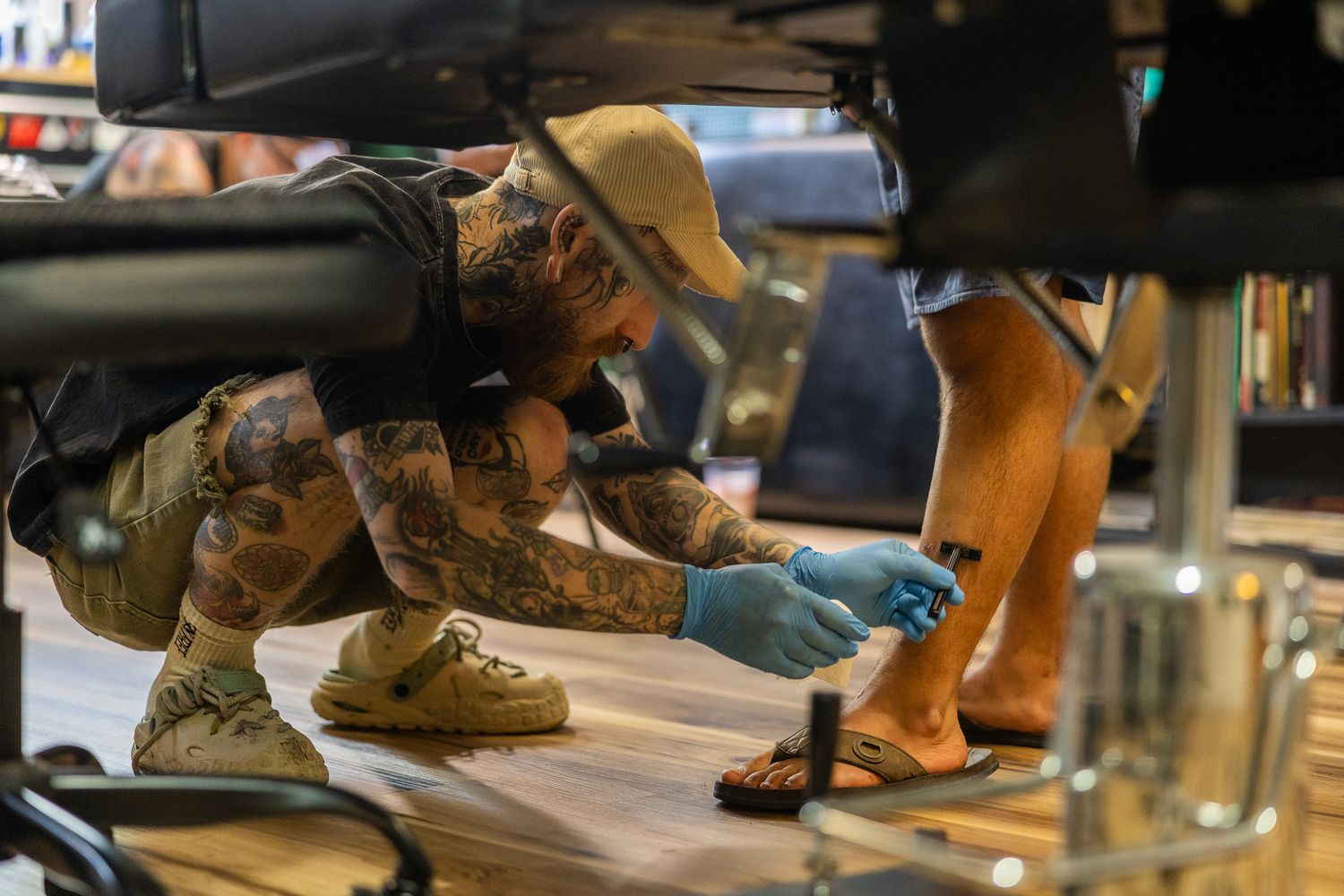 Tattooed artist in blue gloves tattooing a clients foot on the floor of a tattoo shop.