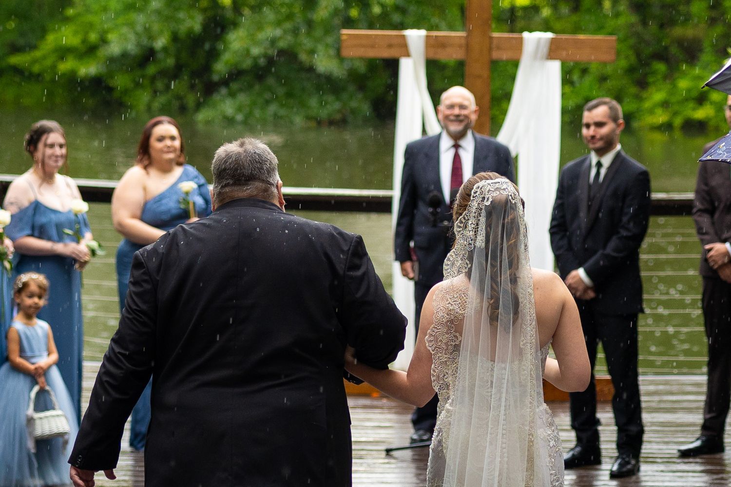Father escorts bride in lace gown toward groom at outdoor rainy wedding ceremony with wooden cross.