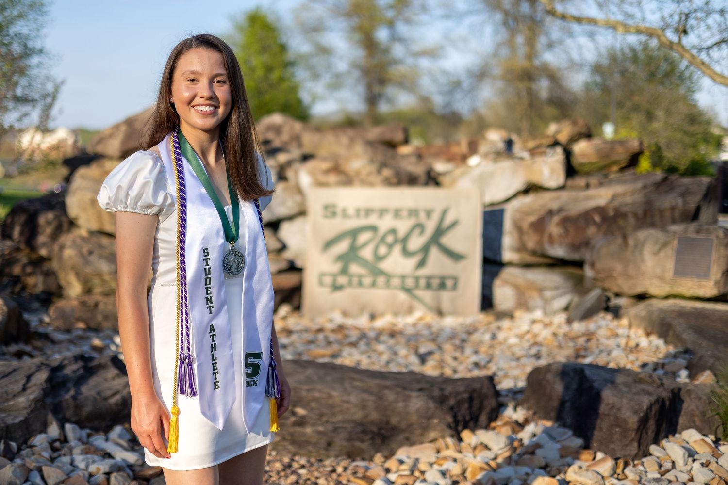 Smiling female graduate in white dress with honor cords poses by Slippery Rock University stone sign.
