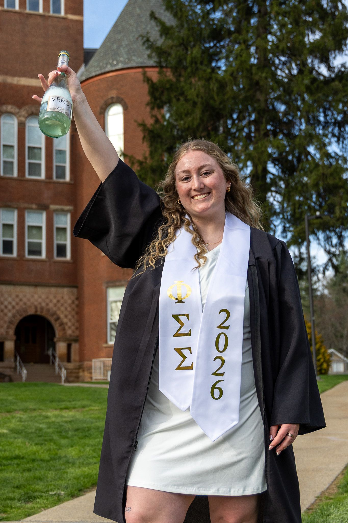 Female graduate in cap and gown raises bottle celebrating with Sigma Sigma 2026 stole.