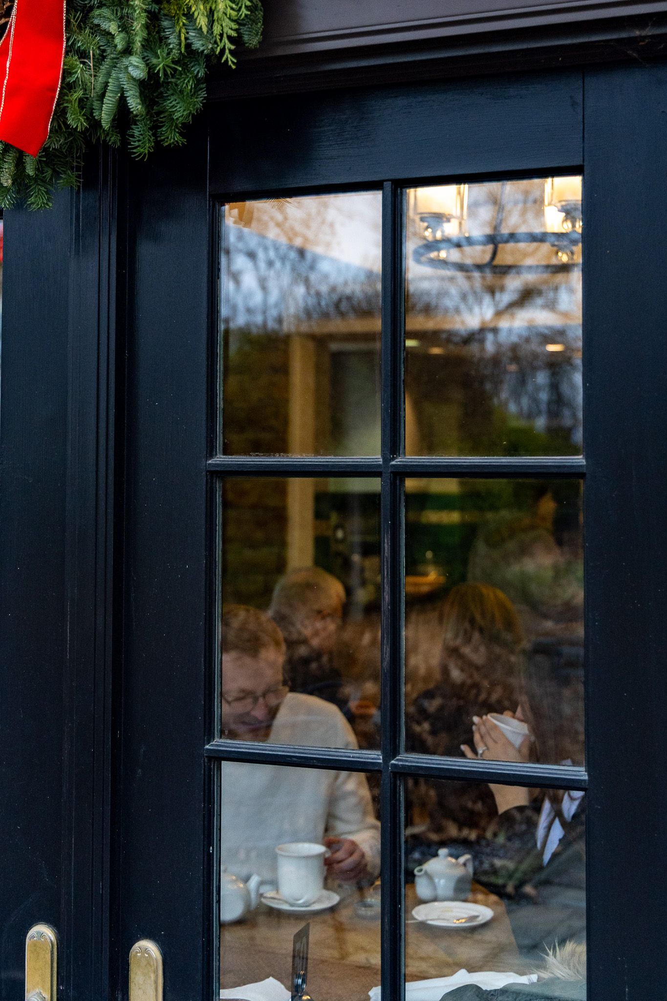 People dining inside a cozy restaurant viewed through a dark-framed window with festive holiday greenery above.