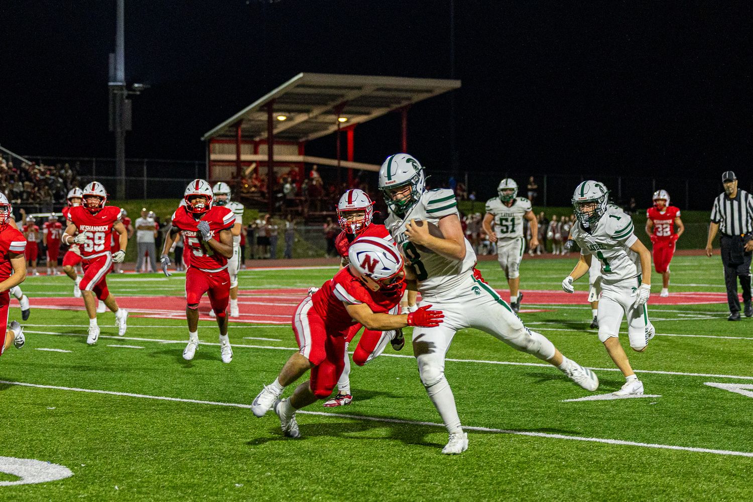 High school football players compete at night, with a green-uniformed player breaking through a tackle attempt by red-uniformed defenders.
