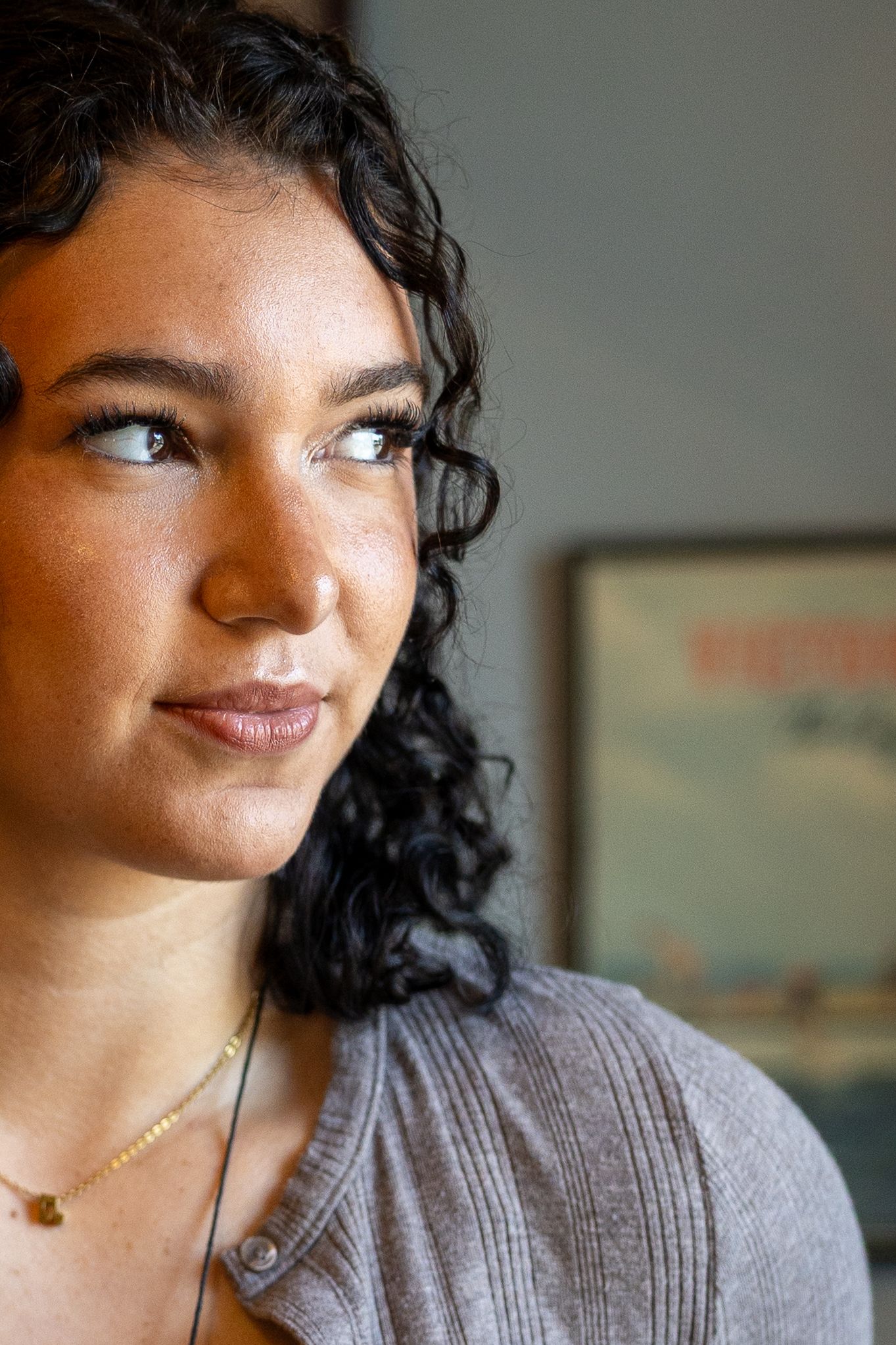 Close-up portrait of a young woman with curly dark hair and light eyes gazing thoughtfully to the side indoors.