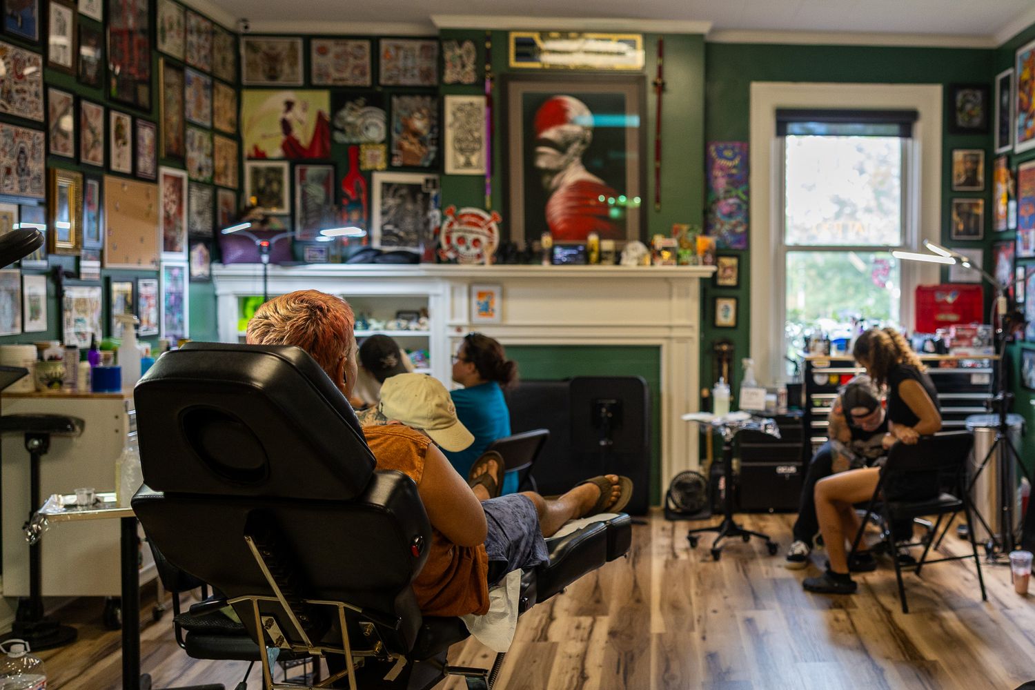 Busy tattoo shop interior with clients getting tattooed, walls covered in artwork, green walls, hardwood floors.