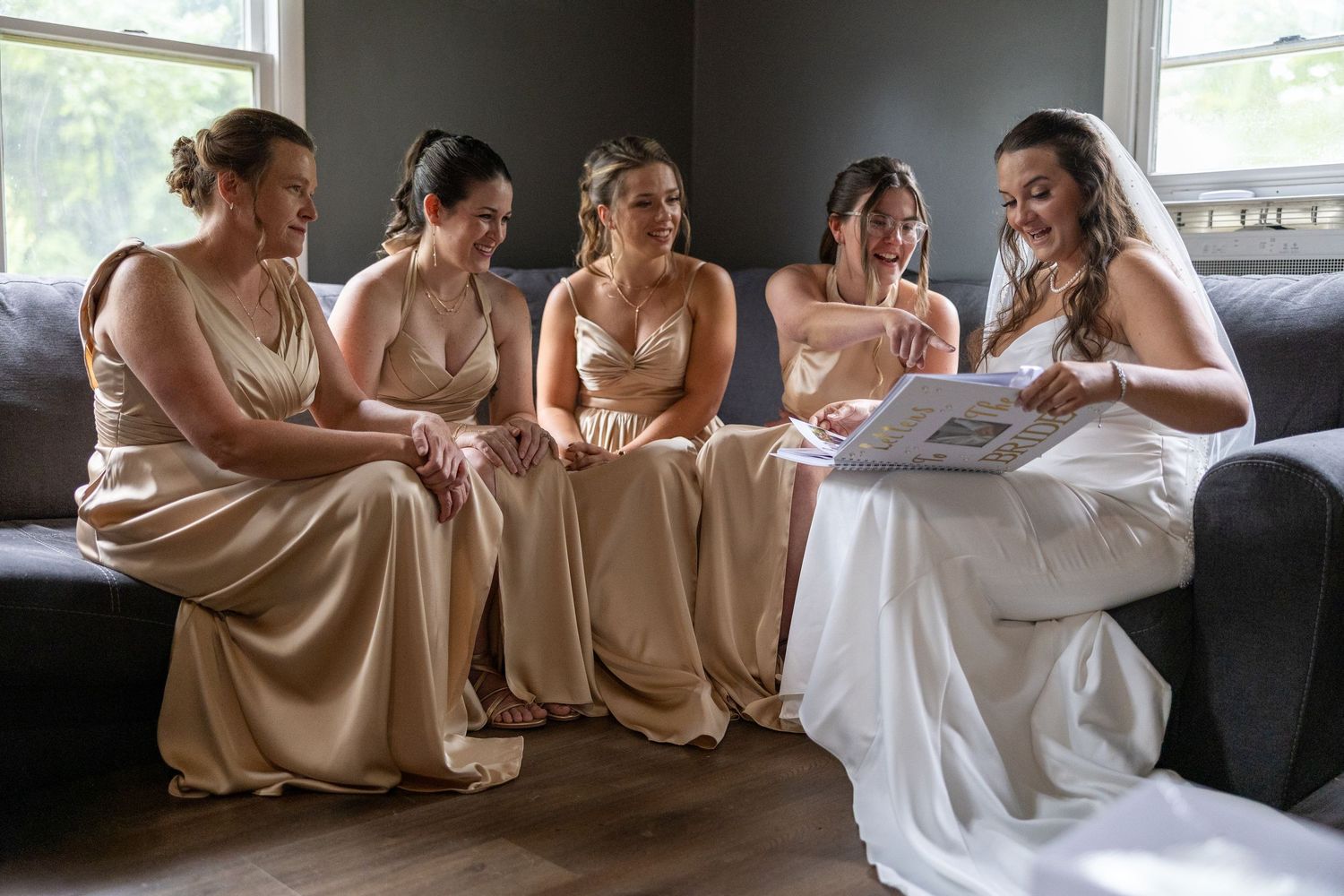 Bride in white gown reads a book with four bridesmaids in champagne dresses seated on a gray sofa.