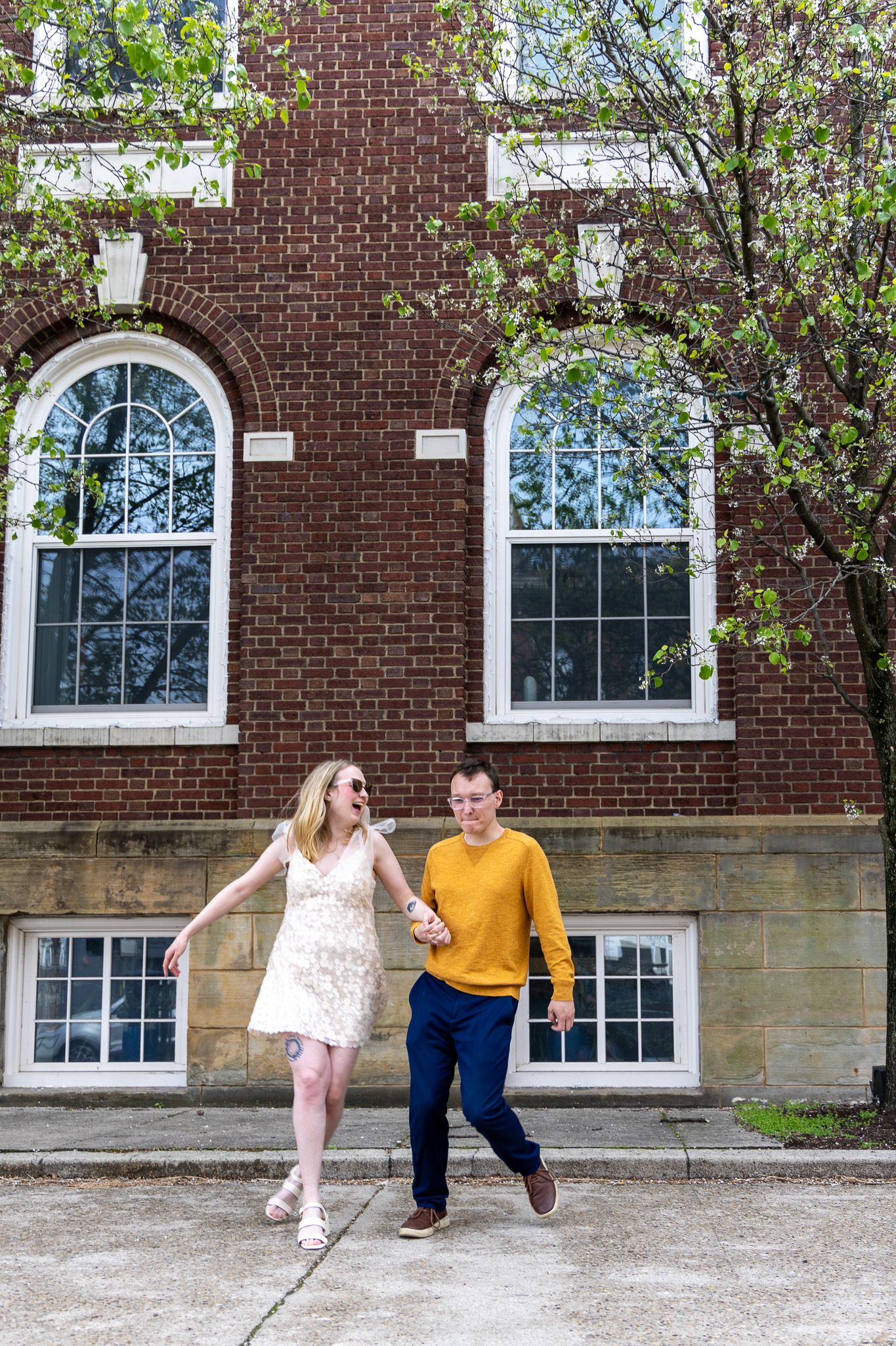 A joyful couple holds hands and walks in front of a red brick building with blooming spring trees.