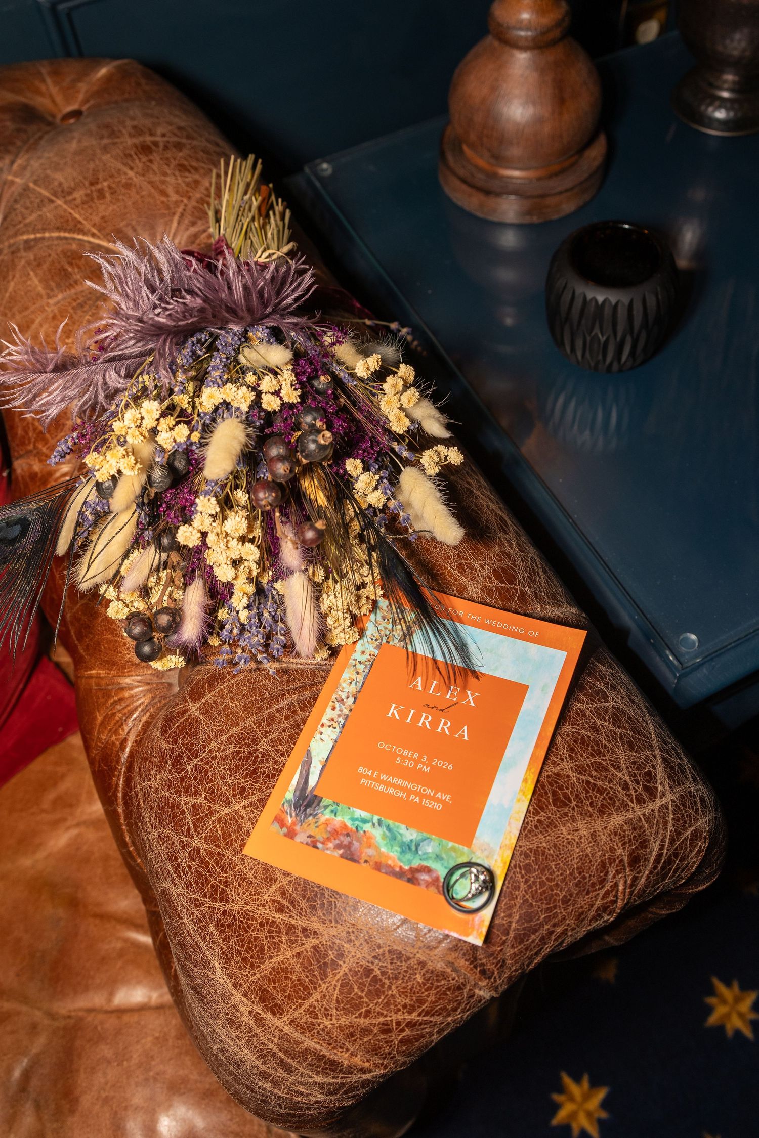Dried floral bouquet beside an orange wedding invitation for Alex and Kirra on a brown leather ottoman.