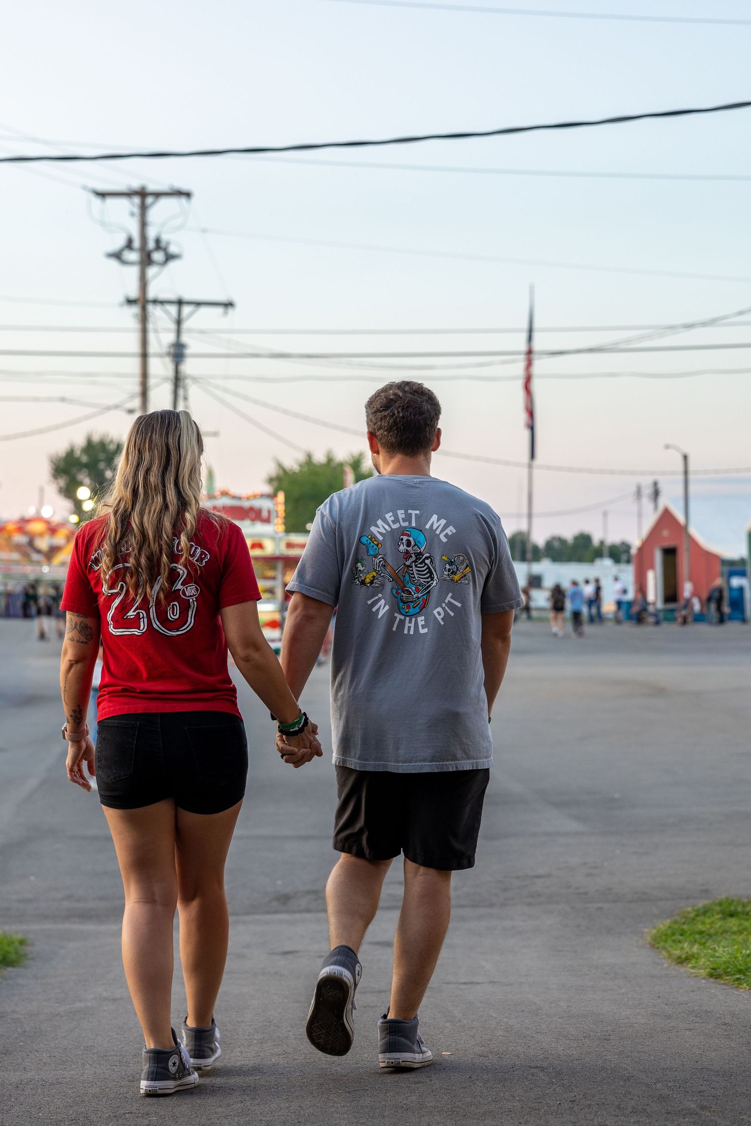 Couple holding hands walking away at outdoor fair, woman in red jersey, man in gray graphic tee, dusk setting.