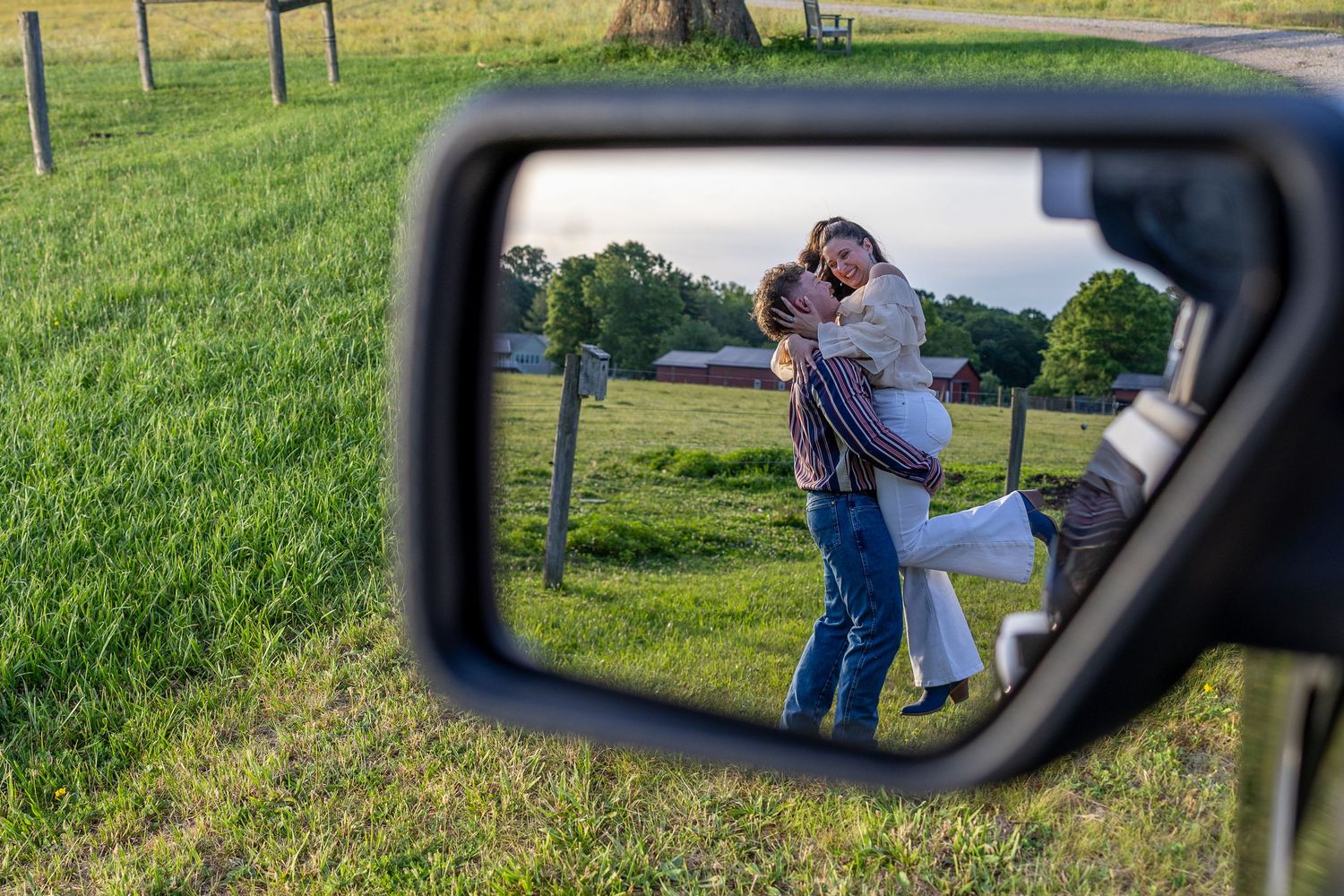 A romantic couple embracing in a green field, captured creatively in a vehicle side mirror reflection.