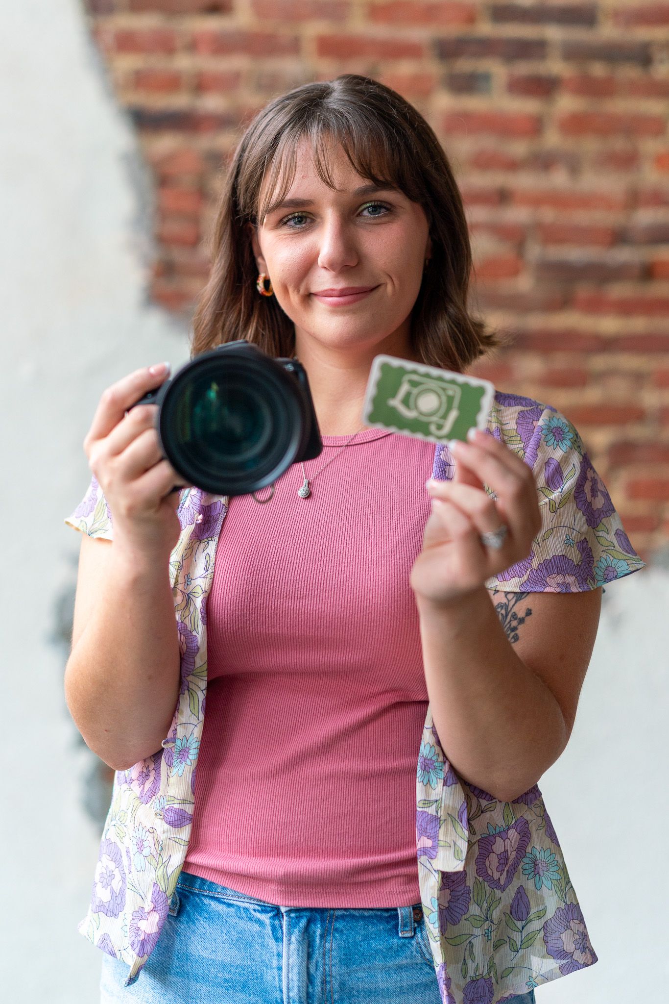 Young woman holding a DSLR camera and a small green card, smiling outdoors near a brick wall.