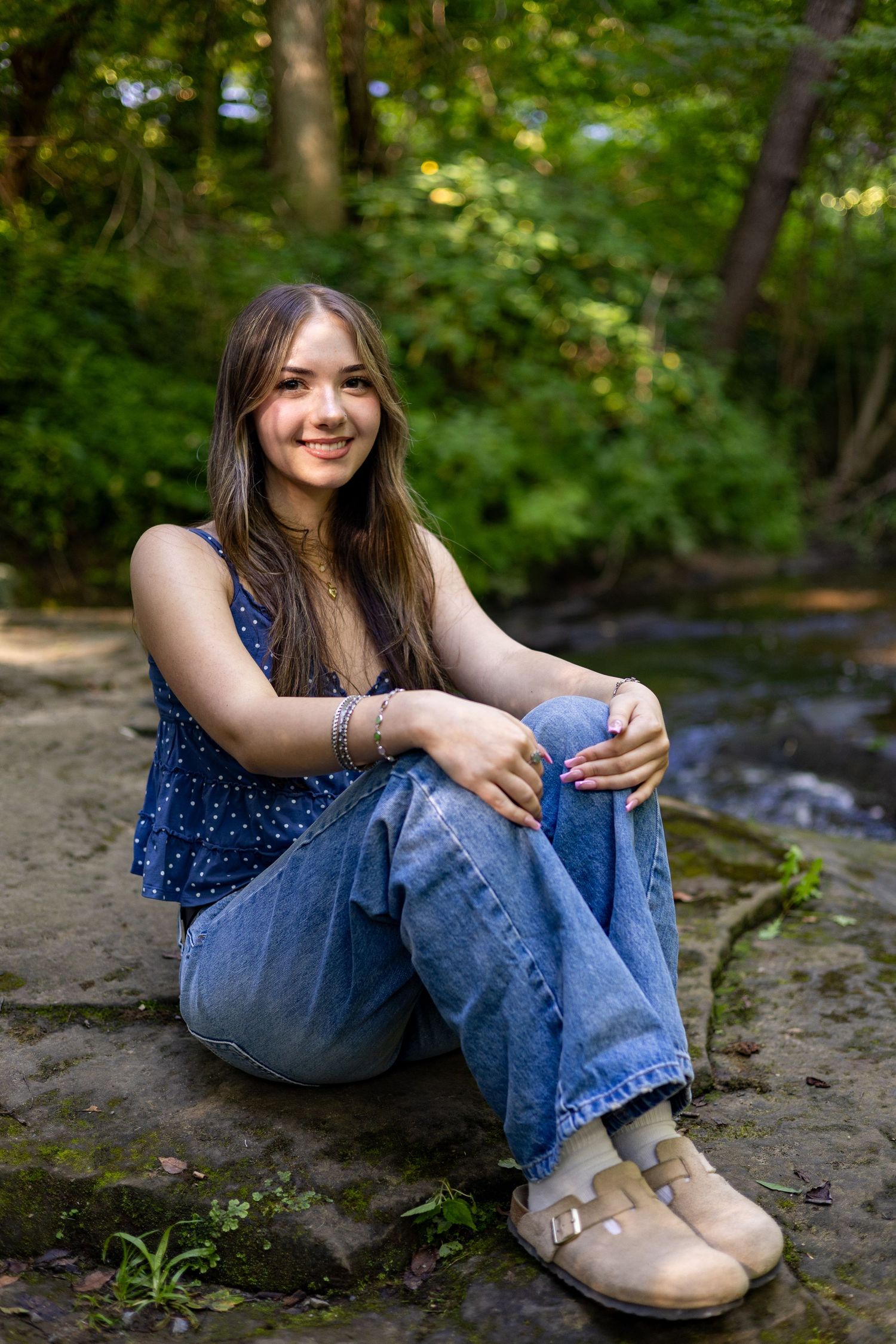 Young woman in blue top and jeans sitting on rocks by a stream in a lush green forest, smiling warmly.