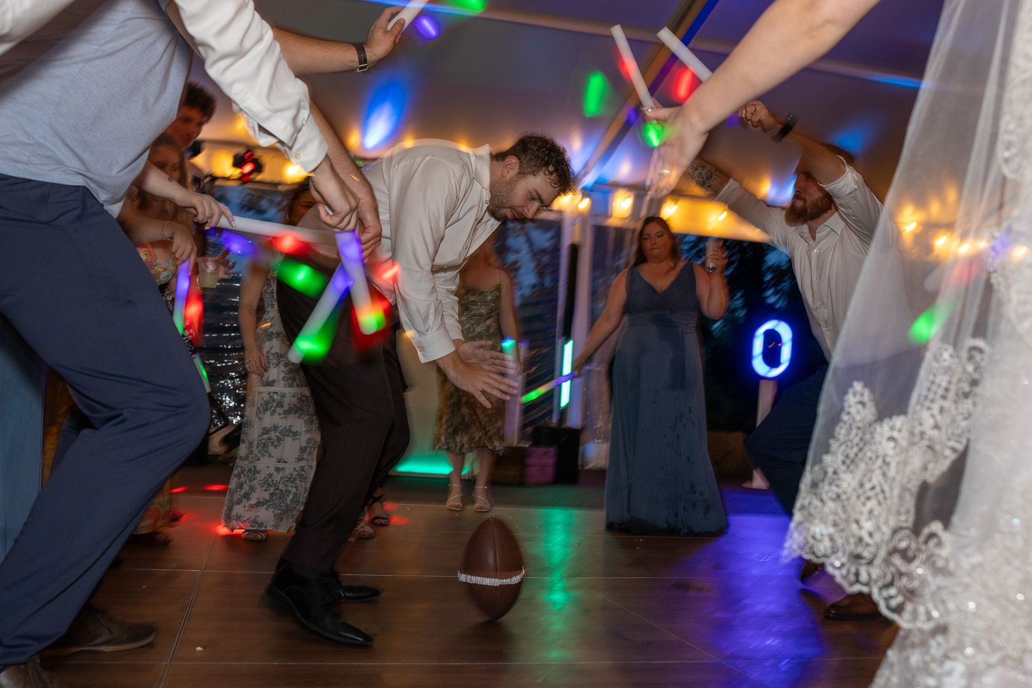 Wedding guests dance energetically with colorful glow sticks on a festive reception dance floor at night.