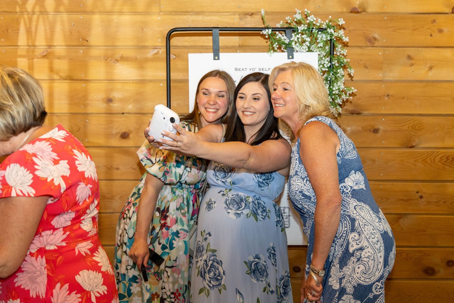 Women in floral dresses taking a selfie with a Polaroid camera at a rustic wooden barn wedding celebration.