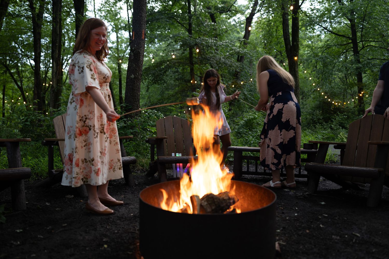 Wedding guests roast marshmallows around a fire pit in a wooded outdoor area with string lights.