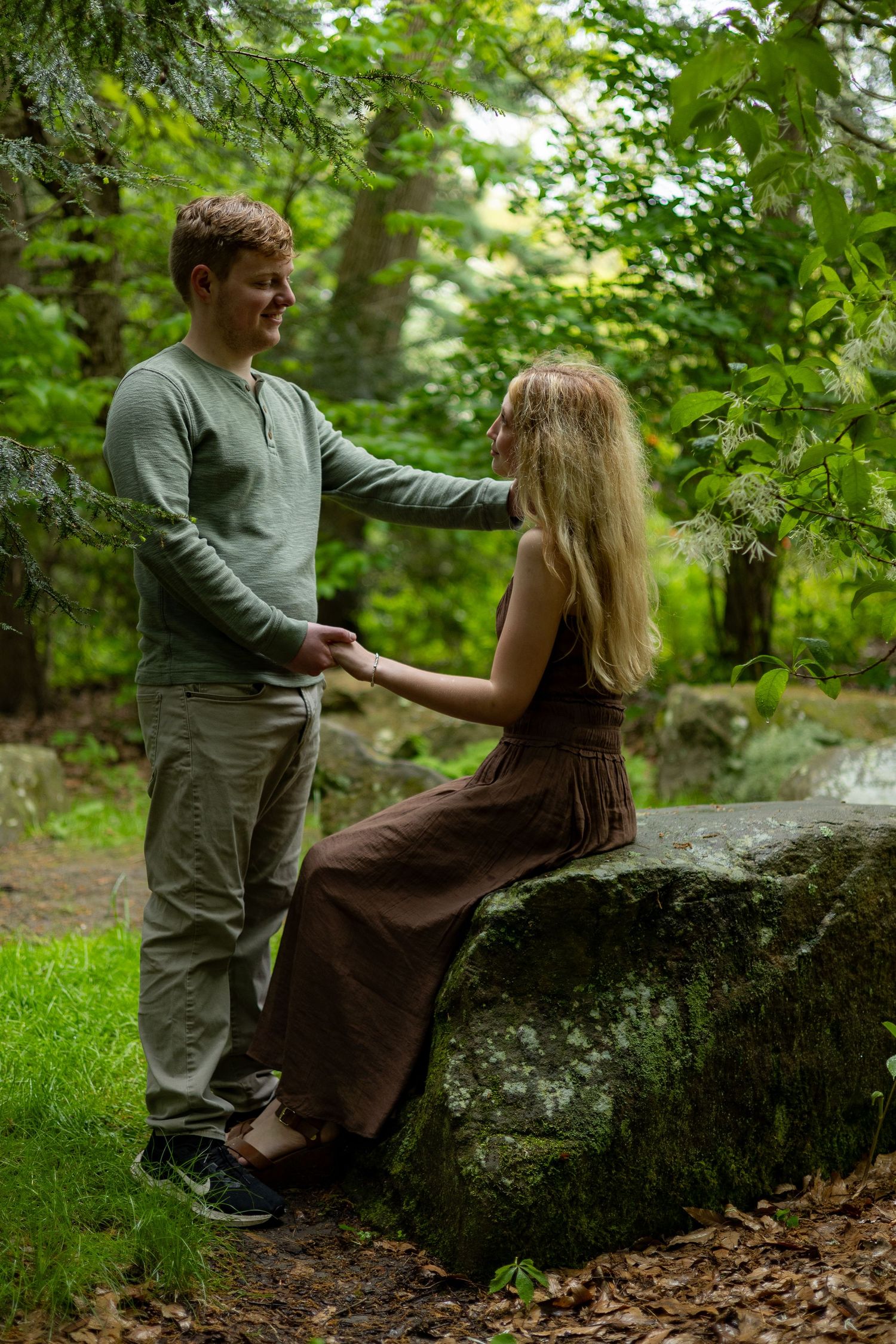 A young couple holds hands in a serene green forest, woman sitting on a mossy rock.